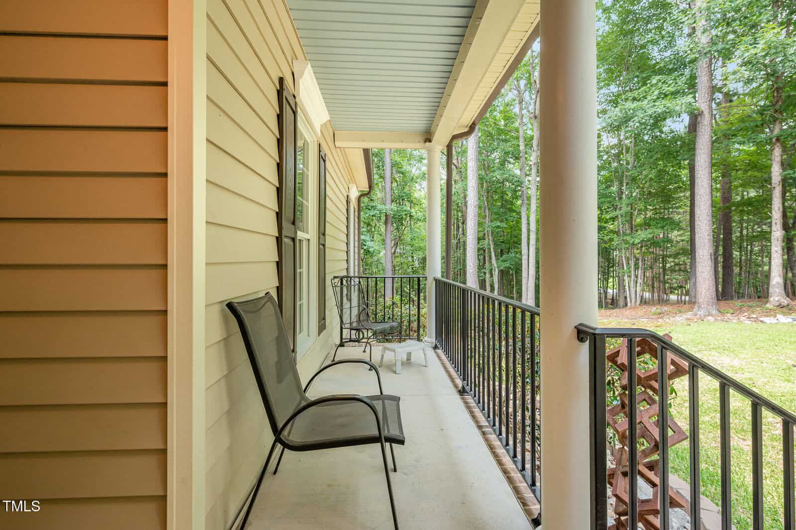 A covered porch with two black metal chairs, a black metal railing, tan siding, and a view of a grassy yard with trees in the background.
