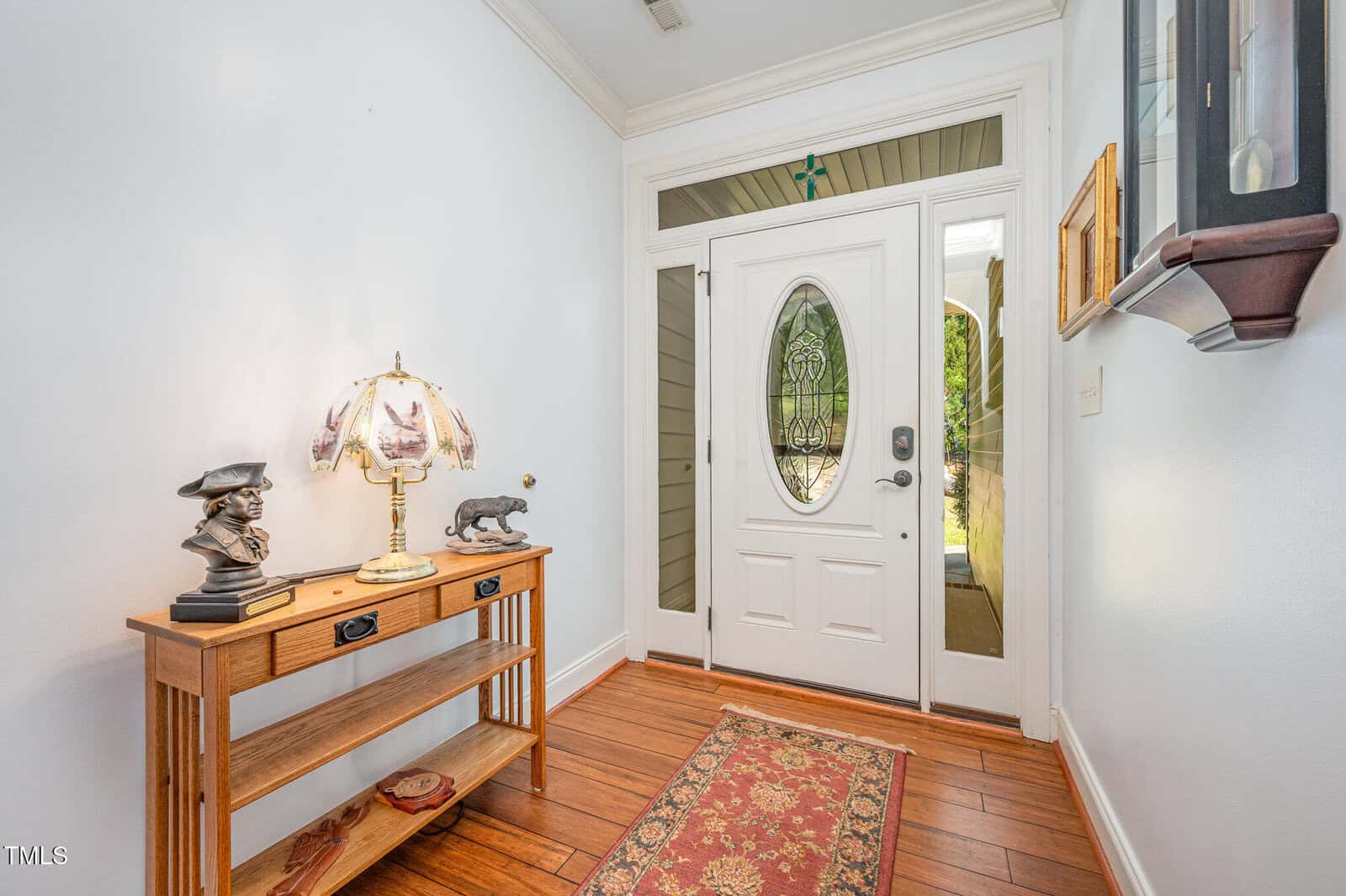 Entryway with a white oval glass front door, light-colored walls, hardwood floor, a small wood table with decorative items, a patterned rug, and a wall clock. Natural light comes through the glass panels.