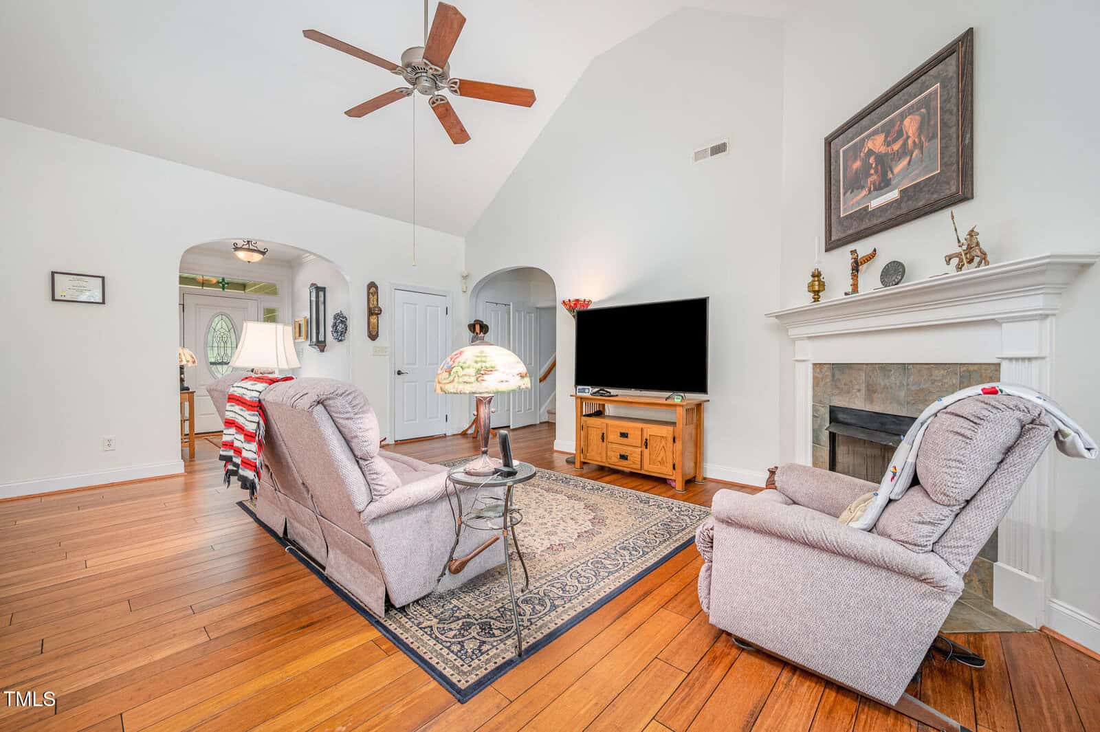 A living room with wood flooring, two recliners facing a TV on a wooden stand, a rug, a fireplace with decor above it, a ceiling fan, and a lamp on a side table. An arched entryway leads to another room.