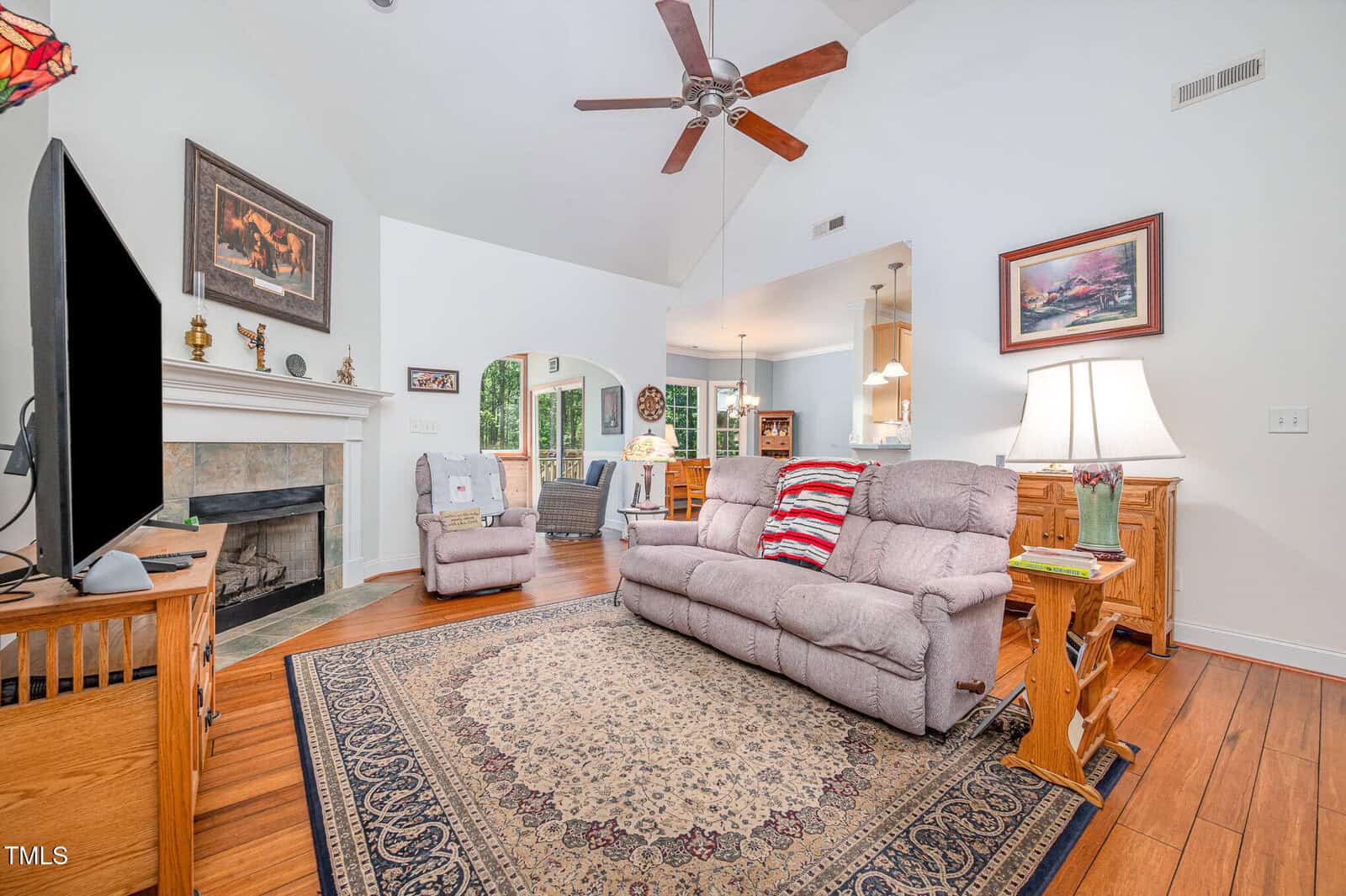 A living room with a beige couch, patterned rug, TV on a wooden stand, fireplace with mantel, armchair, hardwood floor, and ceiling fan. The space connects to a dining area through an arched doorway.