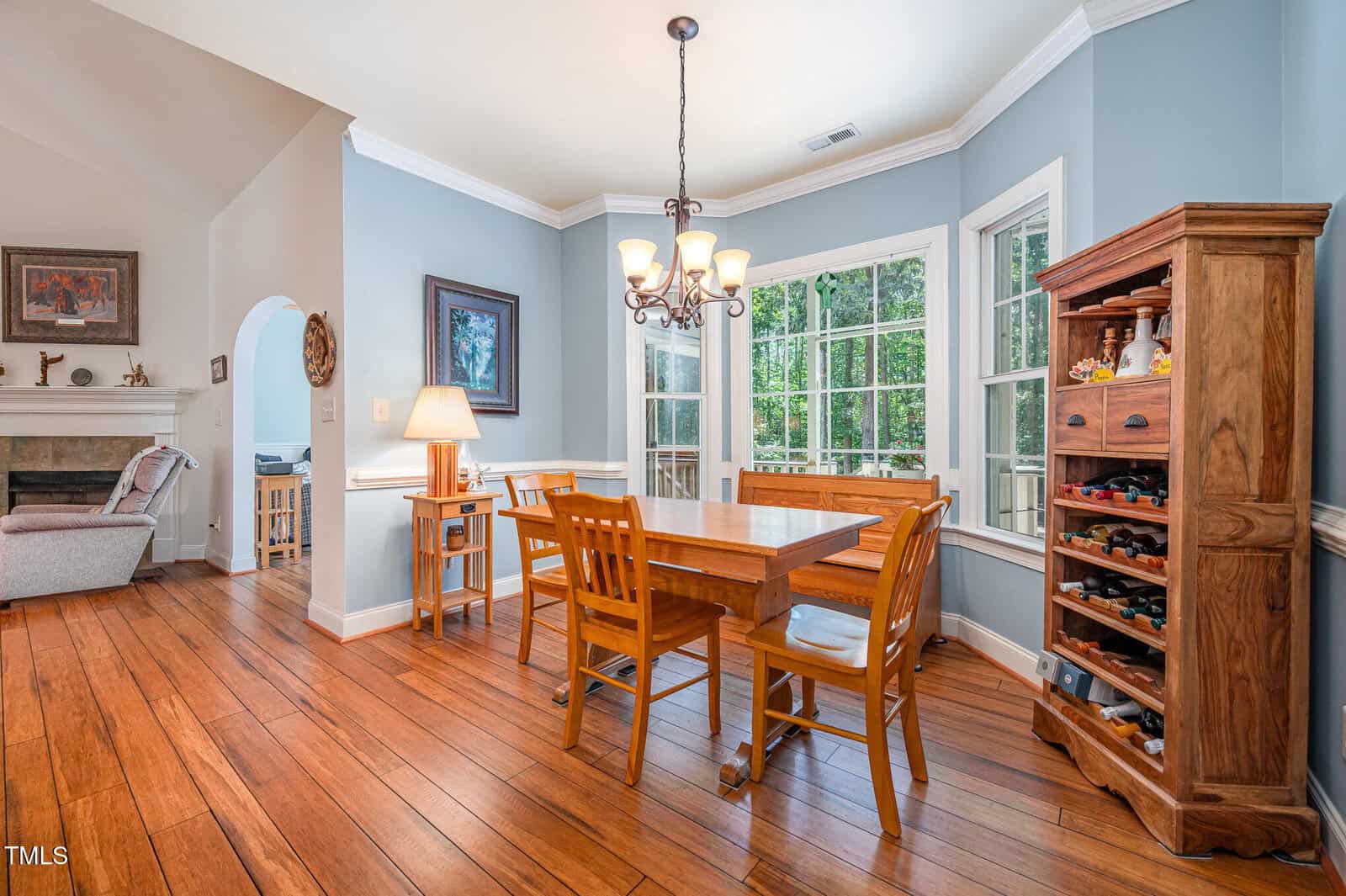 A dining room with wood flooring, a rectangular table and four chairs, a wooden cabinet with wine storage, large windows, a chandelier, and an adjacent living room visible through an arched doorway.