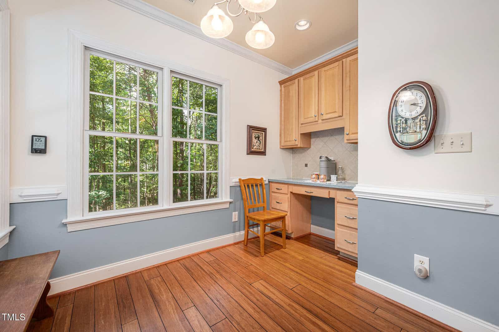 Small home office nook with a wooden desk, chair, and upper cabinets. A clock and light switch are mounted on the wall. Large window provides a view of trees outside. Hardwood floor and chandelier overhead.