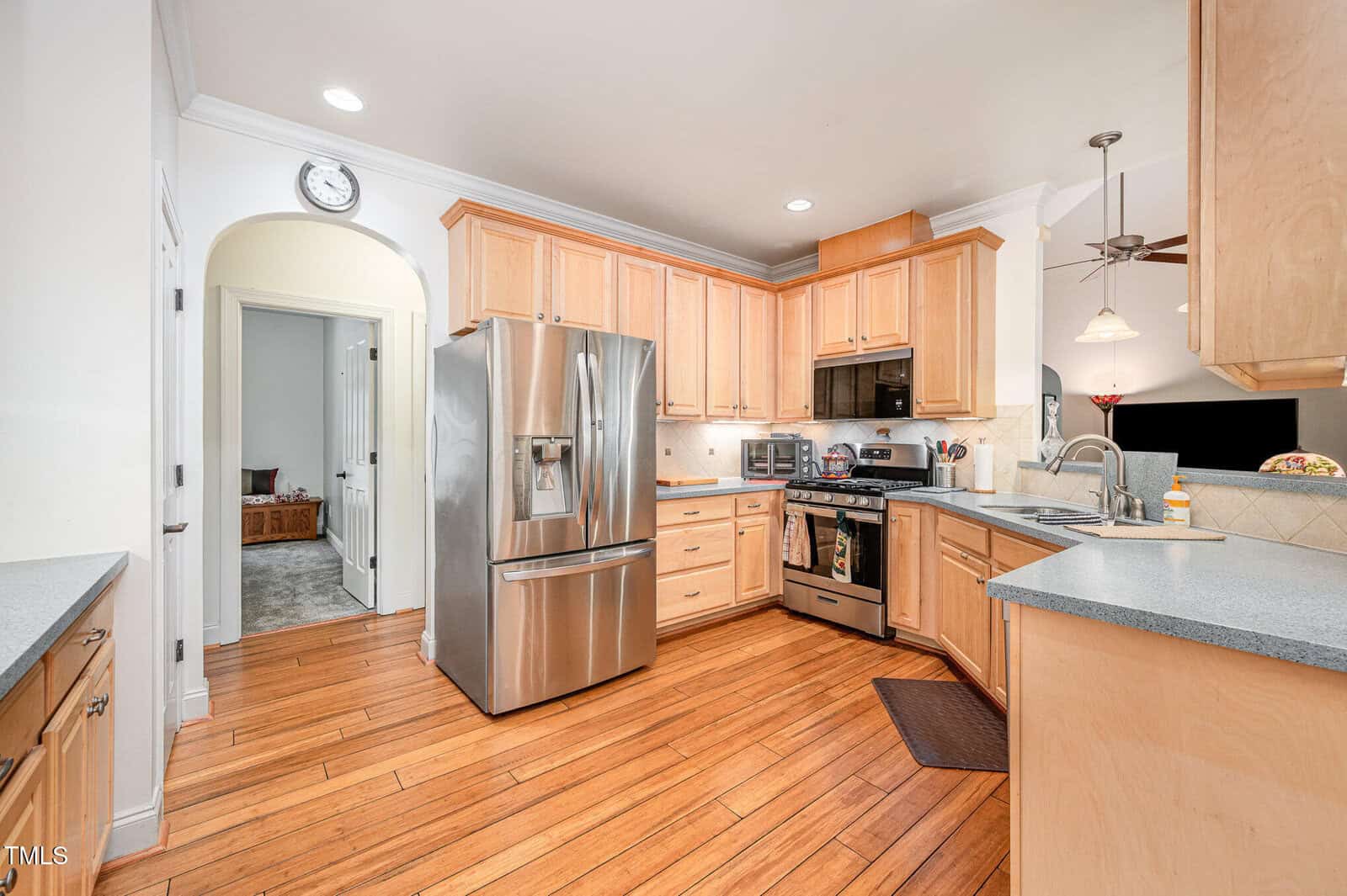 A spacious kitchen with light wood cabinets, stainless steel appliances, a double-door refrigerator, gas stove, microwave, dishwasher, and light wood flooring. Countertops are light grey with various small kitchen items visible.