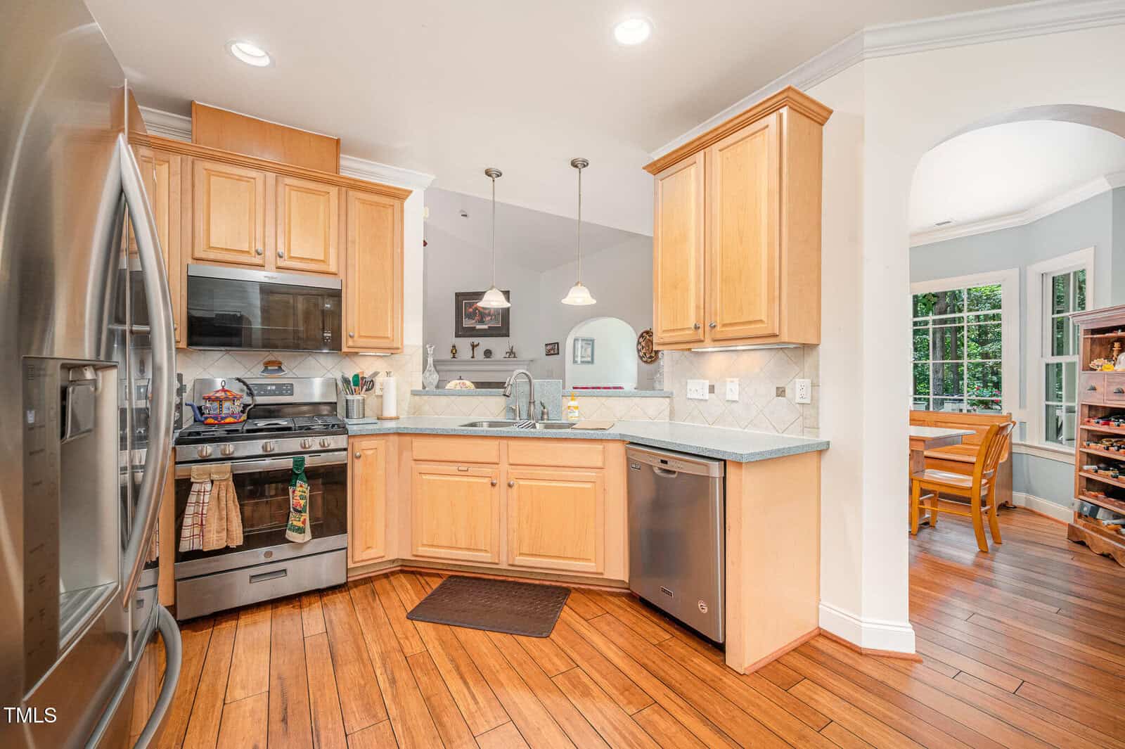 Modern kitchen with stainless steel appliances, wooden cabinets, and hardwood floors. A dining area with a table and chairs is visible through an arched doorway in the background.