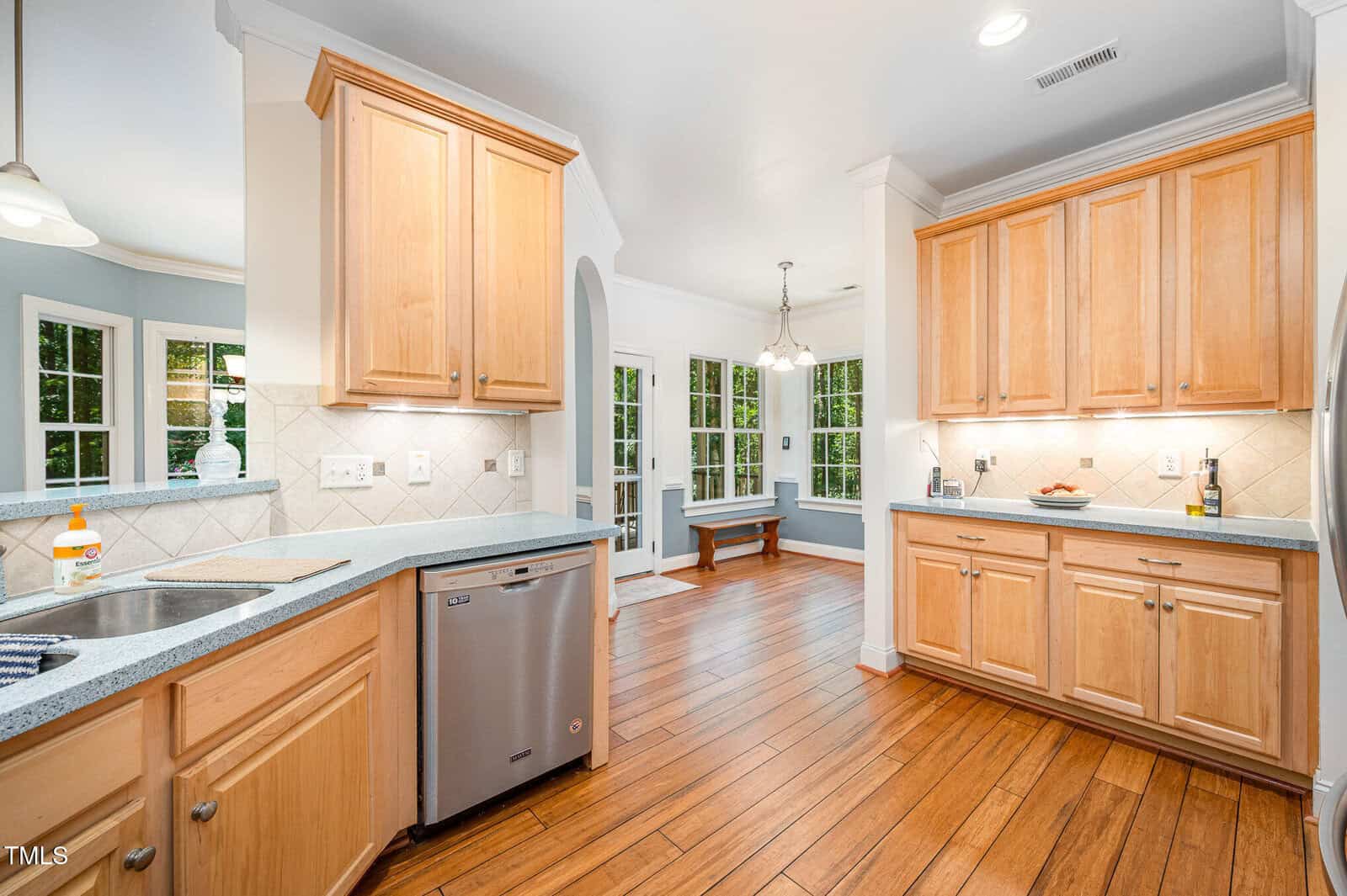 Bright kitchen with wood cabinets, stainless steel dishwasher, and light countertops. The space opens to a dining area with large windows, a bench, and glass doors leading outside. Hardwood floors run throughout.