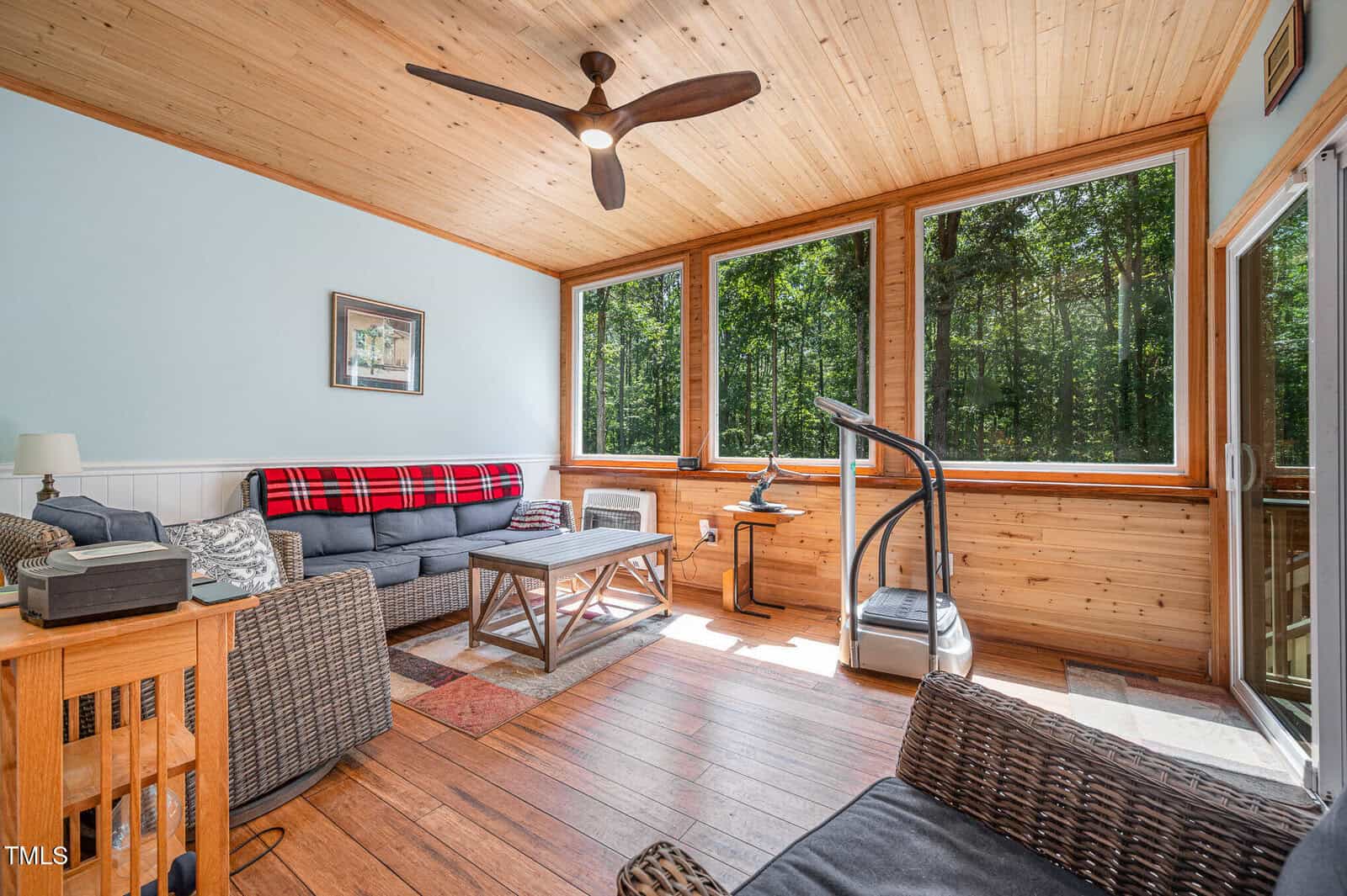 Bright sunroom with wood floors, large windows, and a wood ceiling. Furnished with a sofa, armchairs, coffee table, exercise machine, and side tables. Surrounded by trees visible through the windows.
