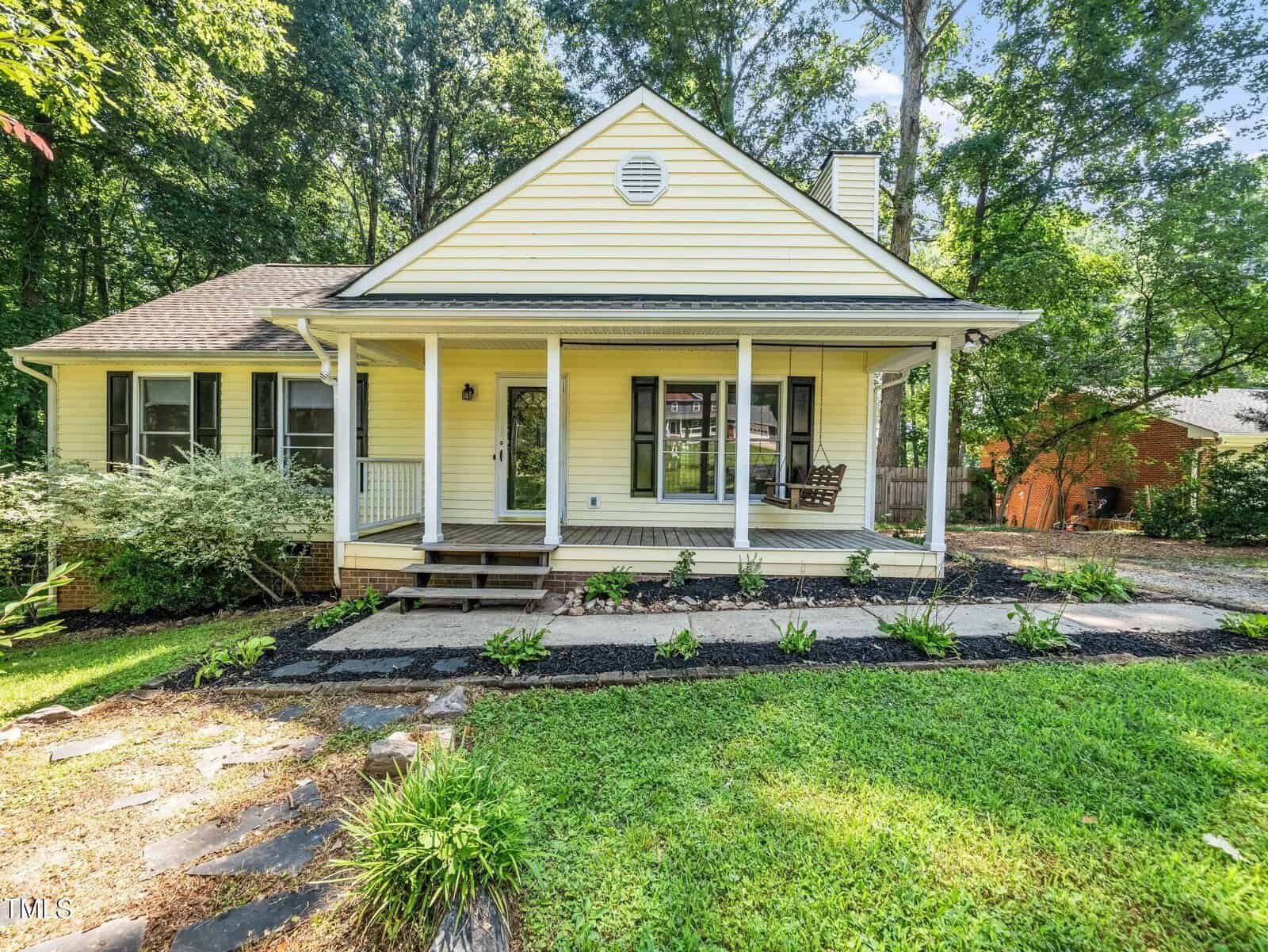 A yellow single-story house with a covered front porch, white trim, and a wooden swing. The yard has green grass, shrubs, and a stone walkway, with tall trees and another house in the background.