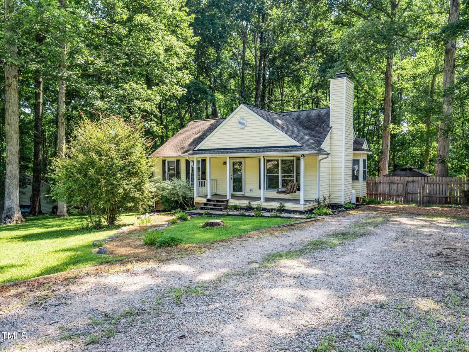 Single-story yellow house with white trim, a covered front porch, and a chimney, surrounded by trees and greenery; gravel driveway leads up to the house under a shaded yard.