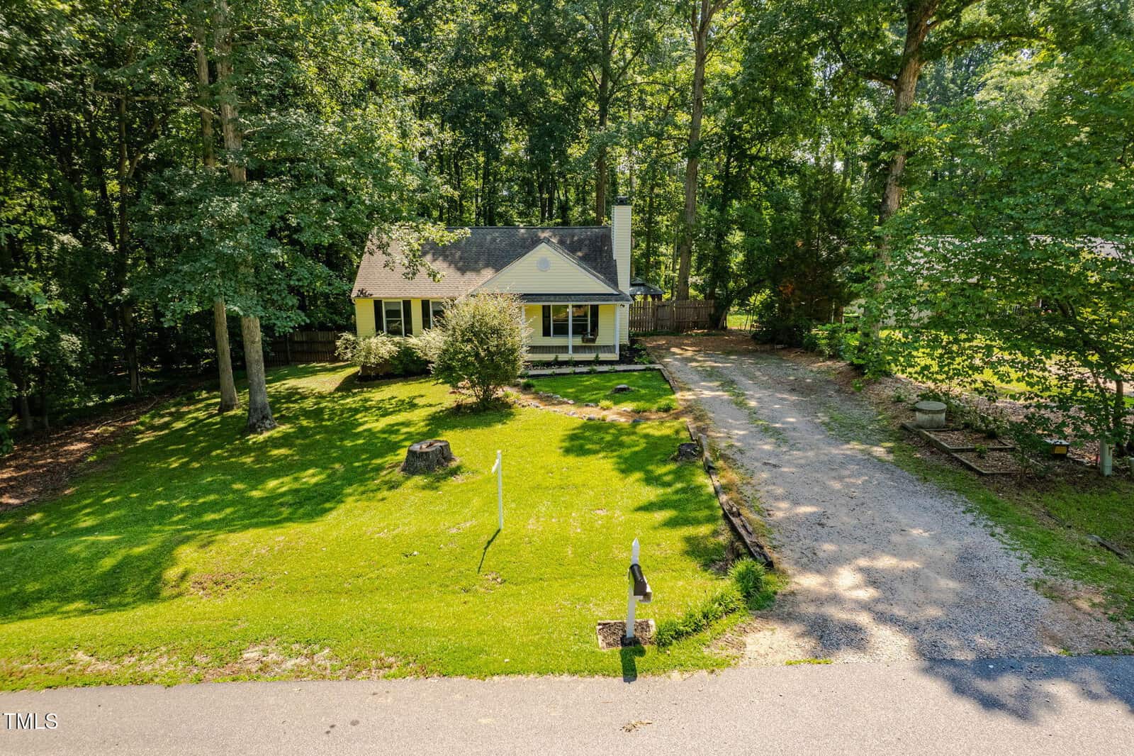 A small yellow house with a front porch sits among tall trees, with a large grassy yard, gravel driveway, mailbox, and shadows from the wooded surroundings on a sunny day.