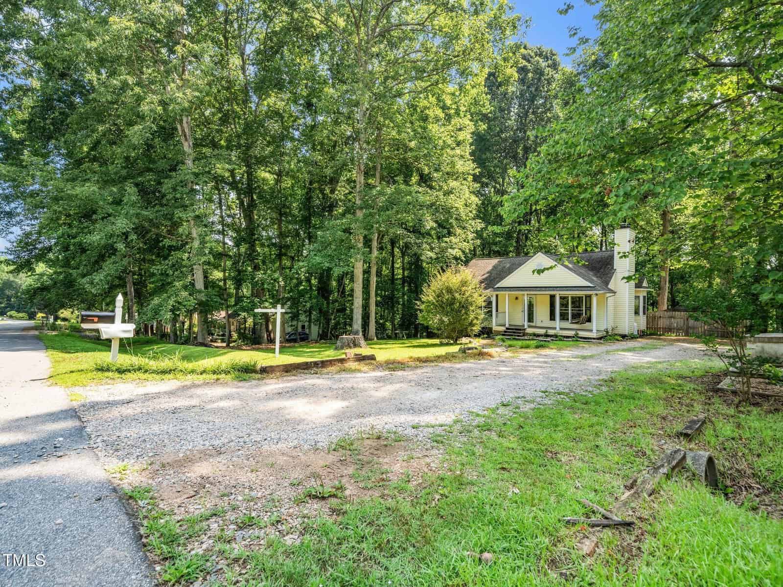 A yellow house with a covered front porch sits among tall trees, facing a gravel driveway and a quiet road. Lush green grass and a white mailbox are visible in the foreground on a sunny day.