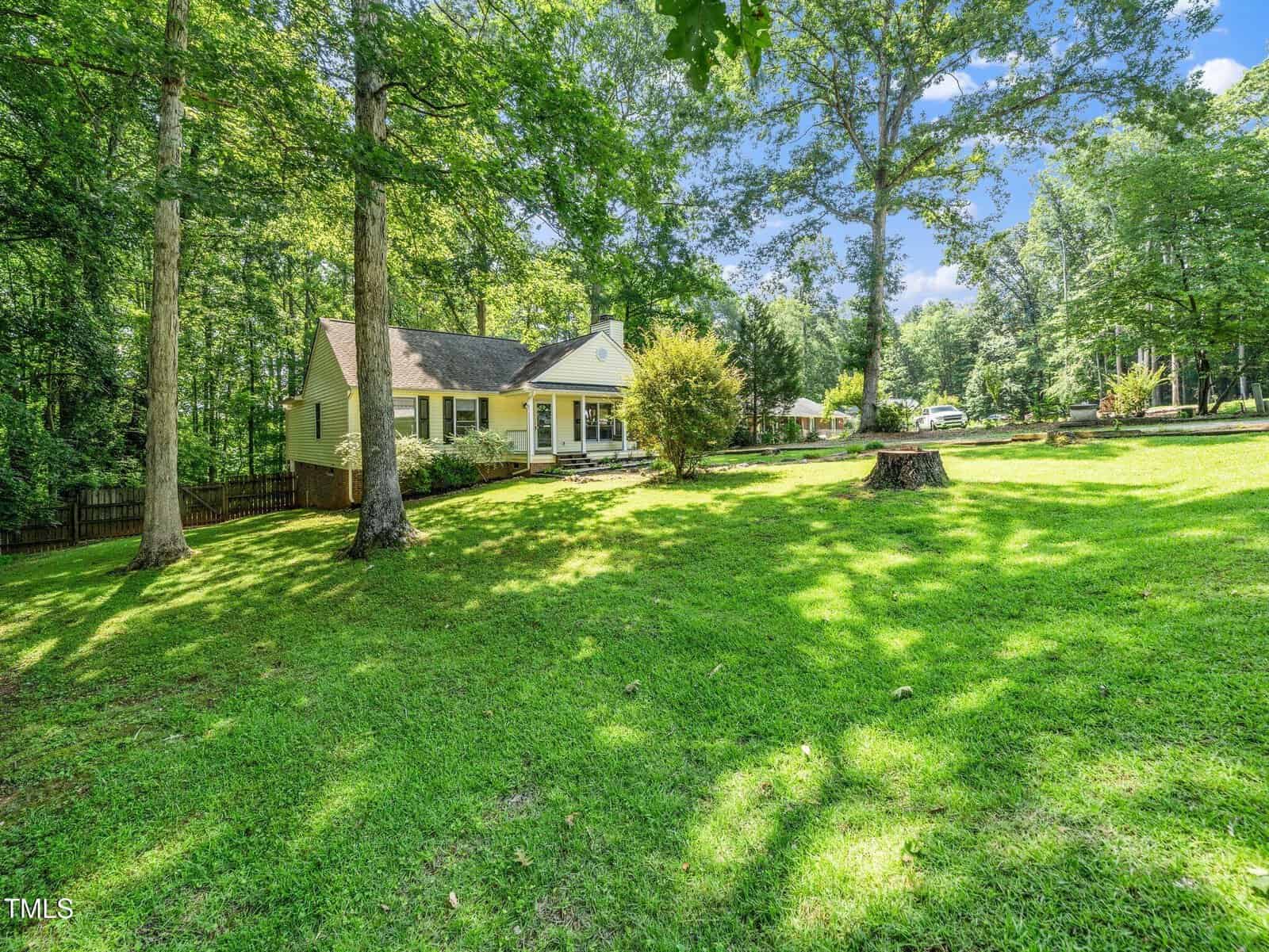 A yellow house with a front porch sits on a large, grassy yard surrounded by tall trees. Sunlight filters through the leaves, and a tree stump is visible in the foreground. The setting is peaceful and suburban.