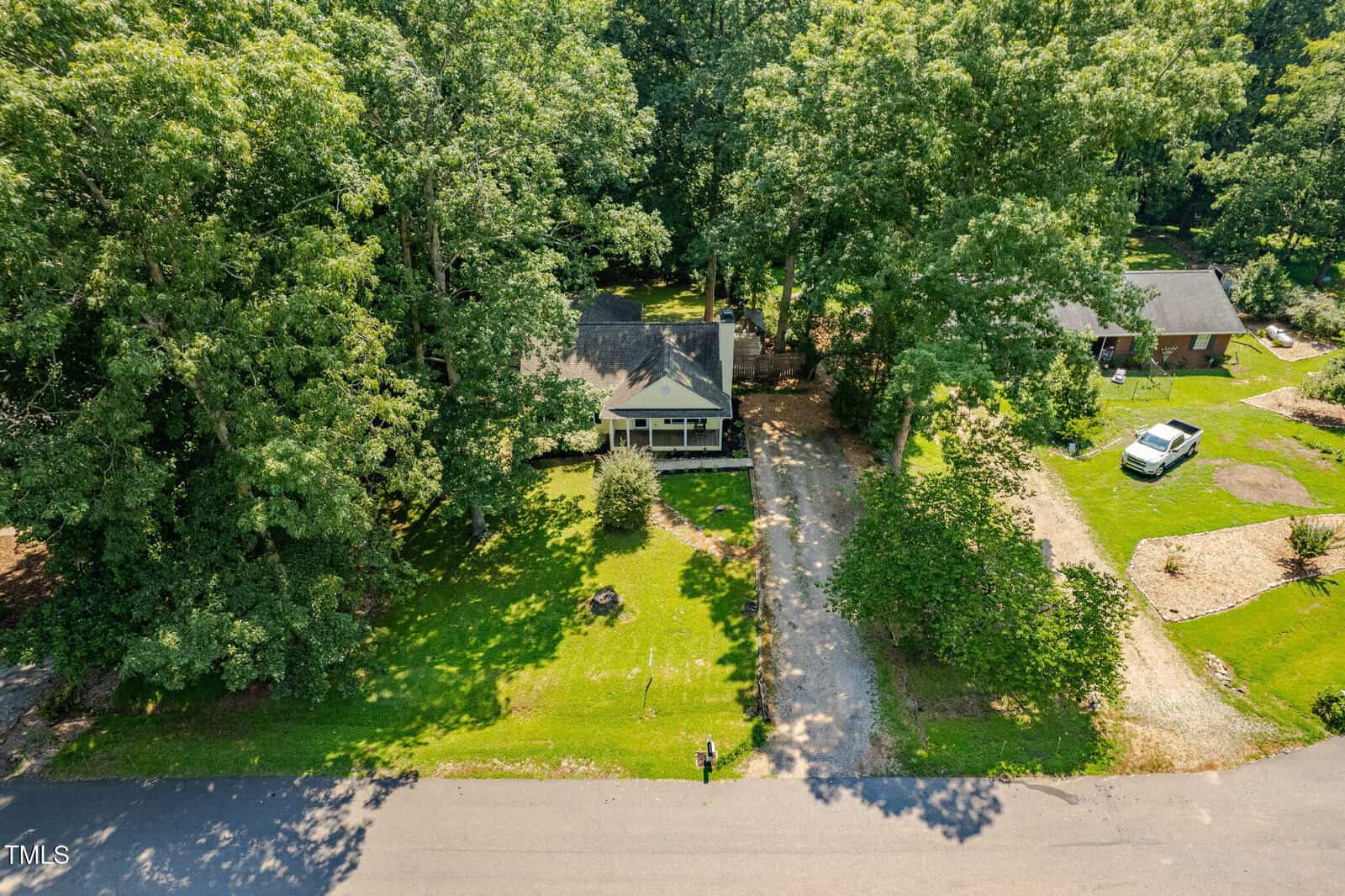 A small house with a covered front porch sits among tall trees with a driveway leading to the street. The lawn is green and well-kept, and a neighboring house with a white car is visible on the right.