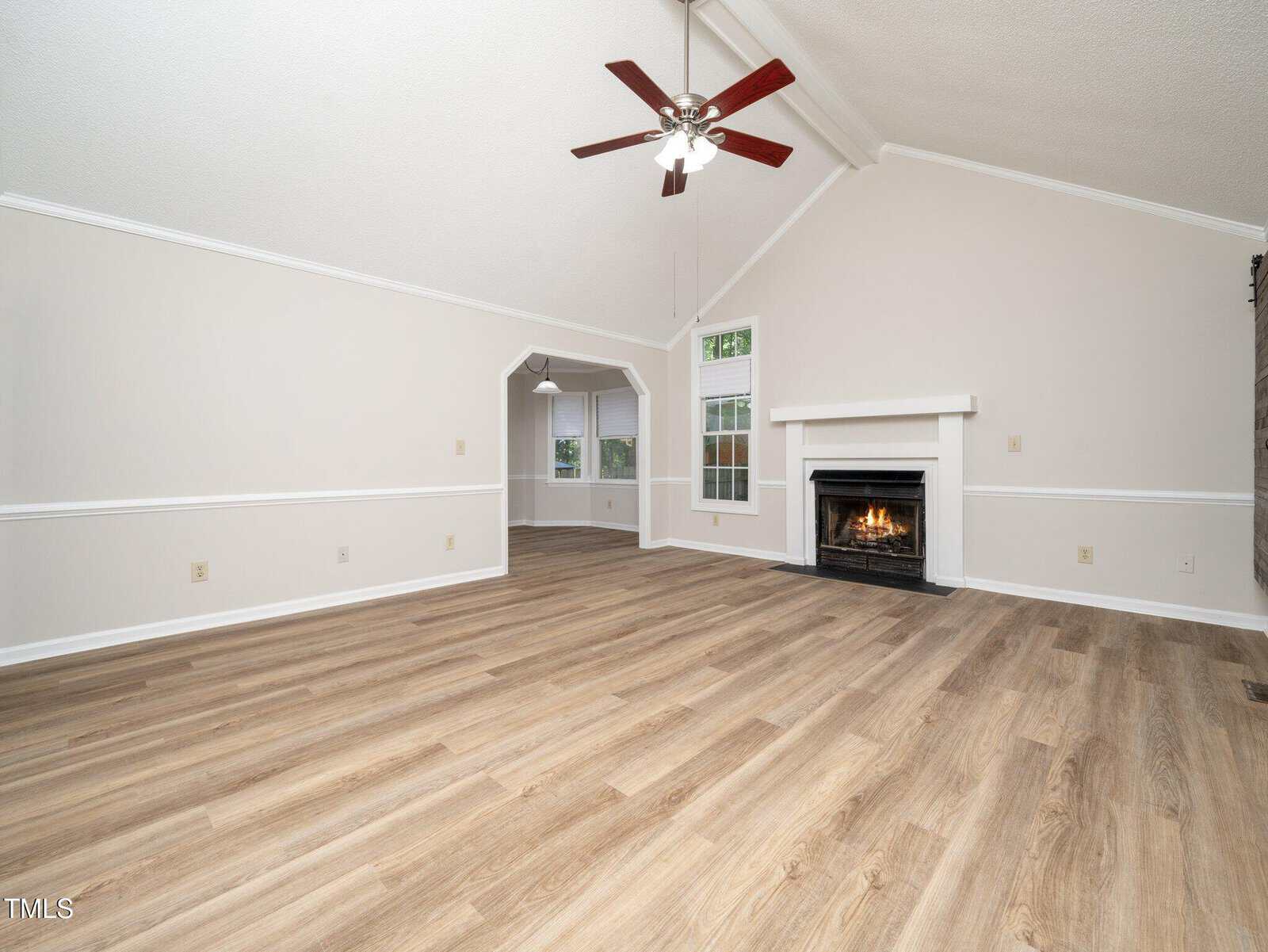 Spacious, empty living room with light wood flooring, a white fireplace, ceiling fan with lights, and an adjacent dining area with windows. Walls are light-colored with white trim.