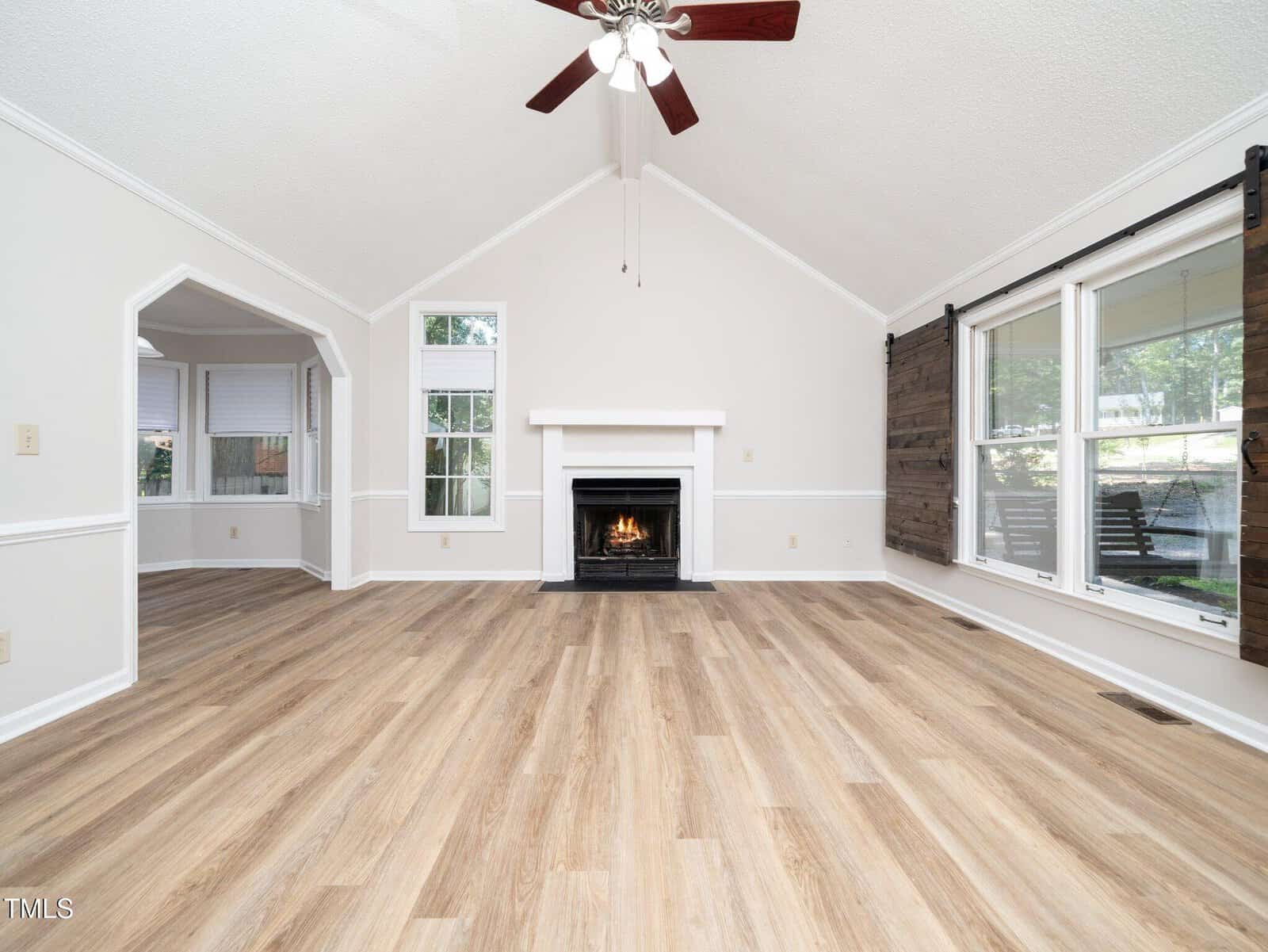 Spacious living room with light wood floors, white walls, a central fireplace, a ceiling fan, large windows to the right, and an open doorway leading to a bright adjacent room.