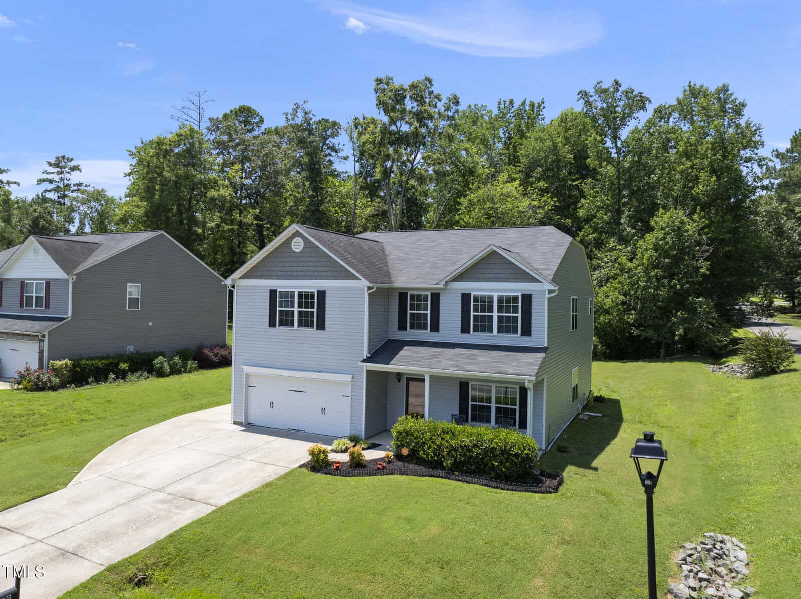 A two-story suburban house with gray siding, white trim, and an attached garage. The home is surrounded by a green lawn, landscaped shrubs, and bordered by trees in the background. A streetlamp stands in the front yard.