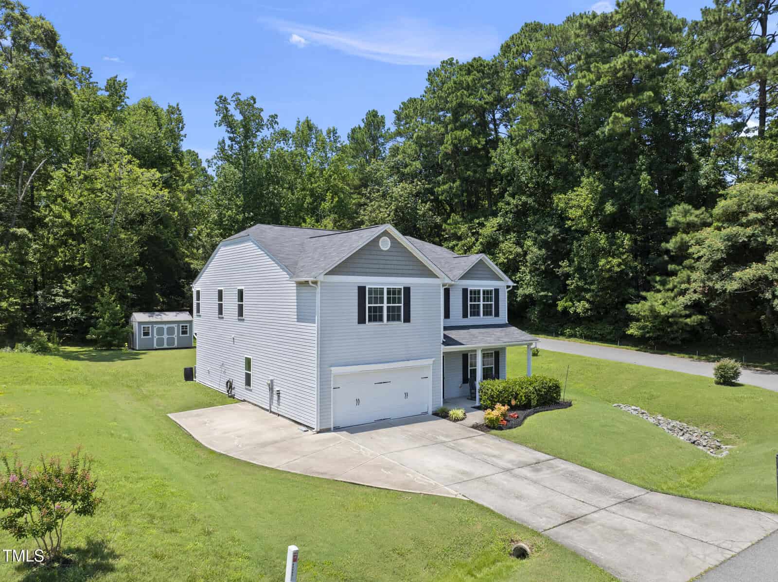 A two-story white house with a double garage sits on a green lawn, bordered by trees. A small shed is visible in the backyard, and the driveway curves toward the street under a clear blue sky.
