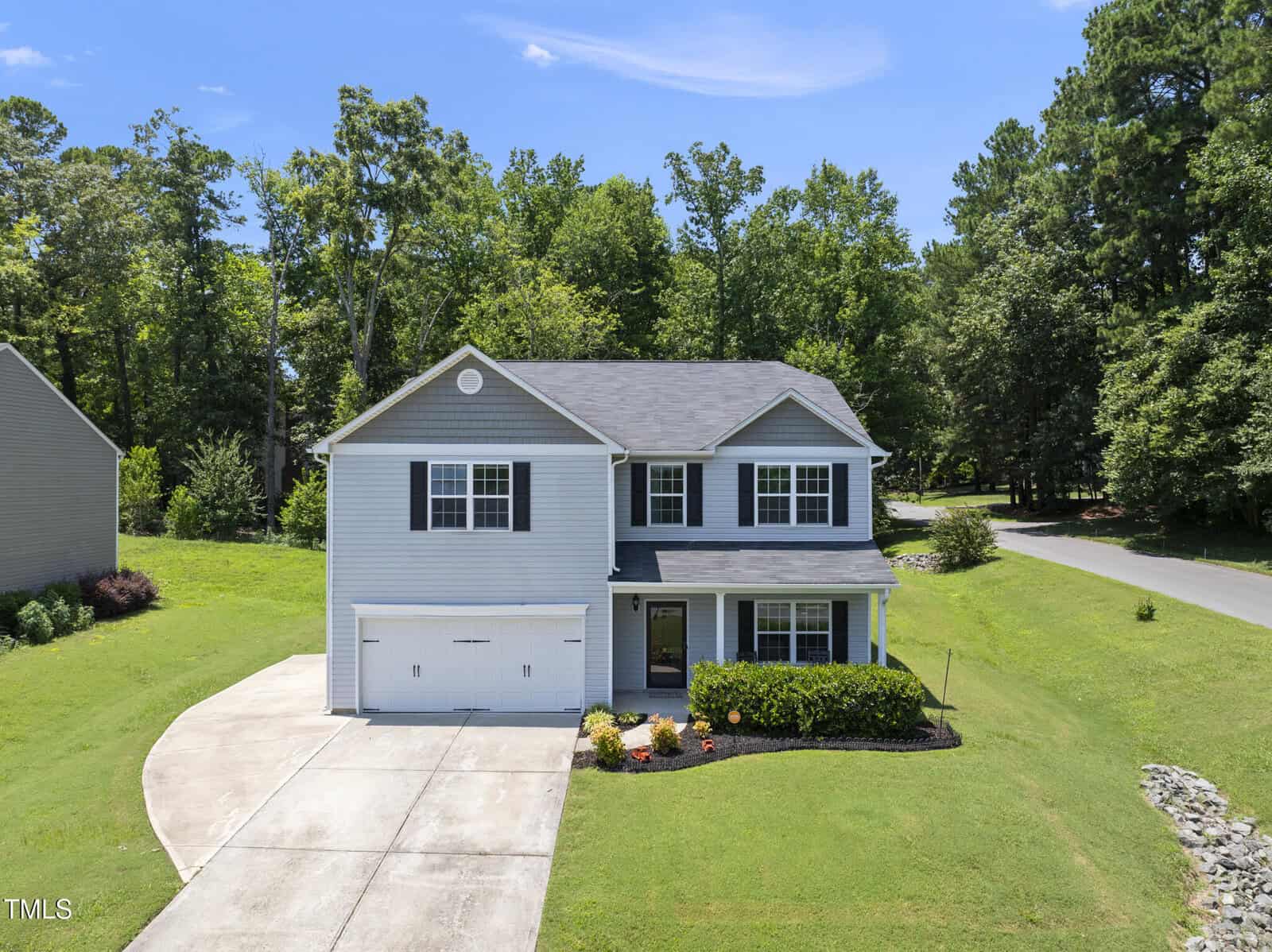 A two-story light gray house with white trim at 702 Pitch Pine Drive in Creedmoor features a double garage, covered front porch, landscaped green lawn, and sits on a corner lot bordered by trees near a wooded area.
