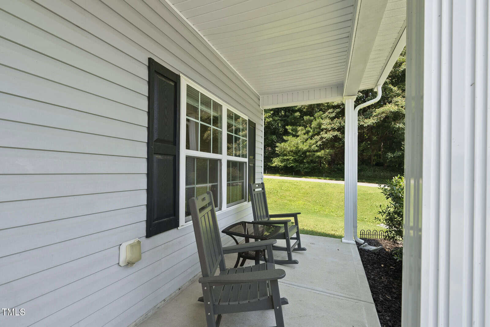 A covered front porch with two black rocking chairs on a concrete floor, white siding, black shutters, and a view of a grassy yard with trees in the background.