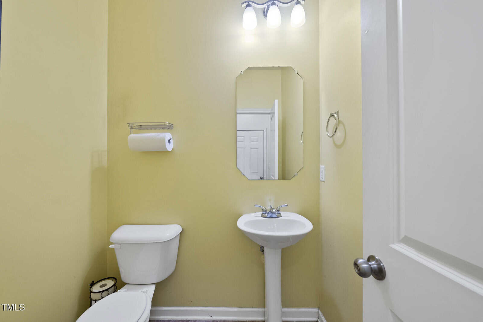 A small bathroom with yellow walls, a toilet, a pedestal sink, an oval mirror, a towel ring, a toilet paper holder, and three lights above the mirror.