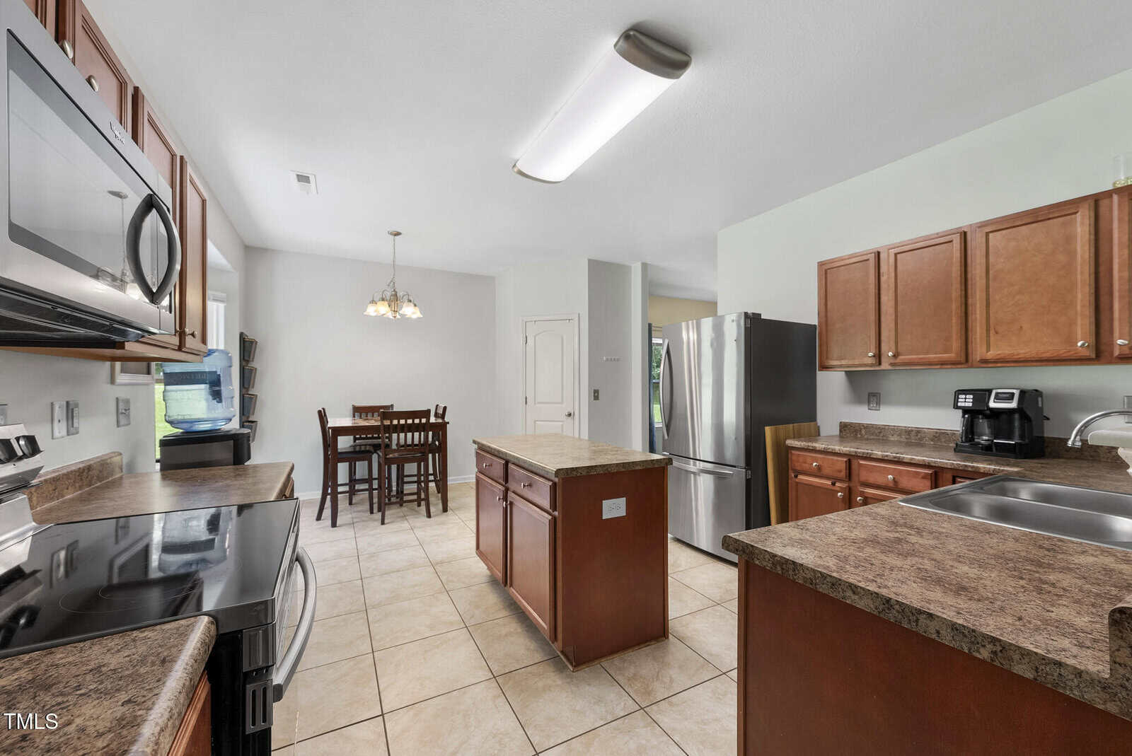 A kitchen with brown cabinets, granite countertops, stainless steel appliances, a central island, tile flooring, and a small dining table with four chairs in a nook by the window.