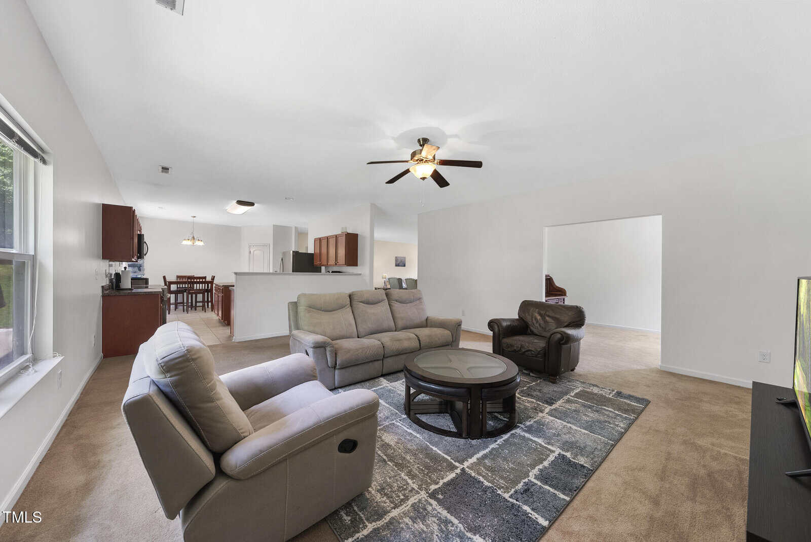 Spacious living room with beige carpet, gray sectional sofa, armchair, round coffee table, TV, ceiling fan, and open view into a kitchen and dining area with wooden cabinets. Large window brings in natural light.