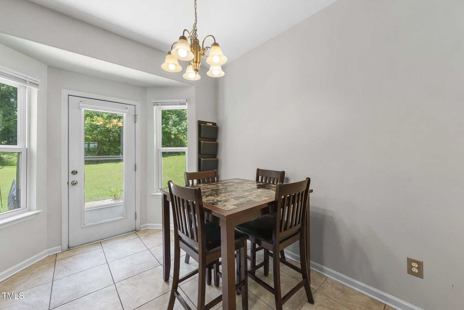A dining area with a high square table and four chairs near windows and a glass door, overlooking a green backyard. A small corner shelf stands against the wall. Light fixture hangs above the table.