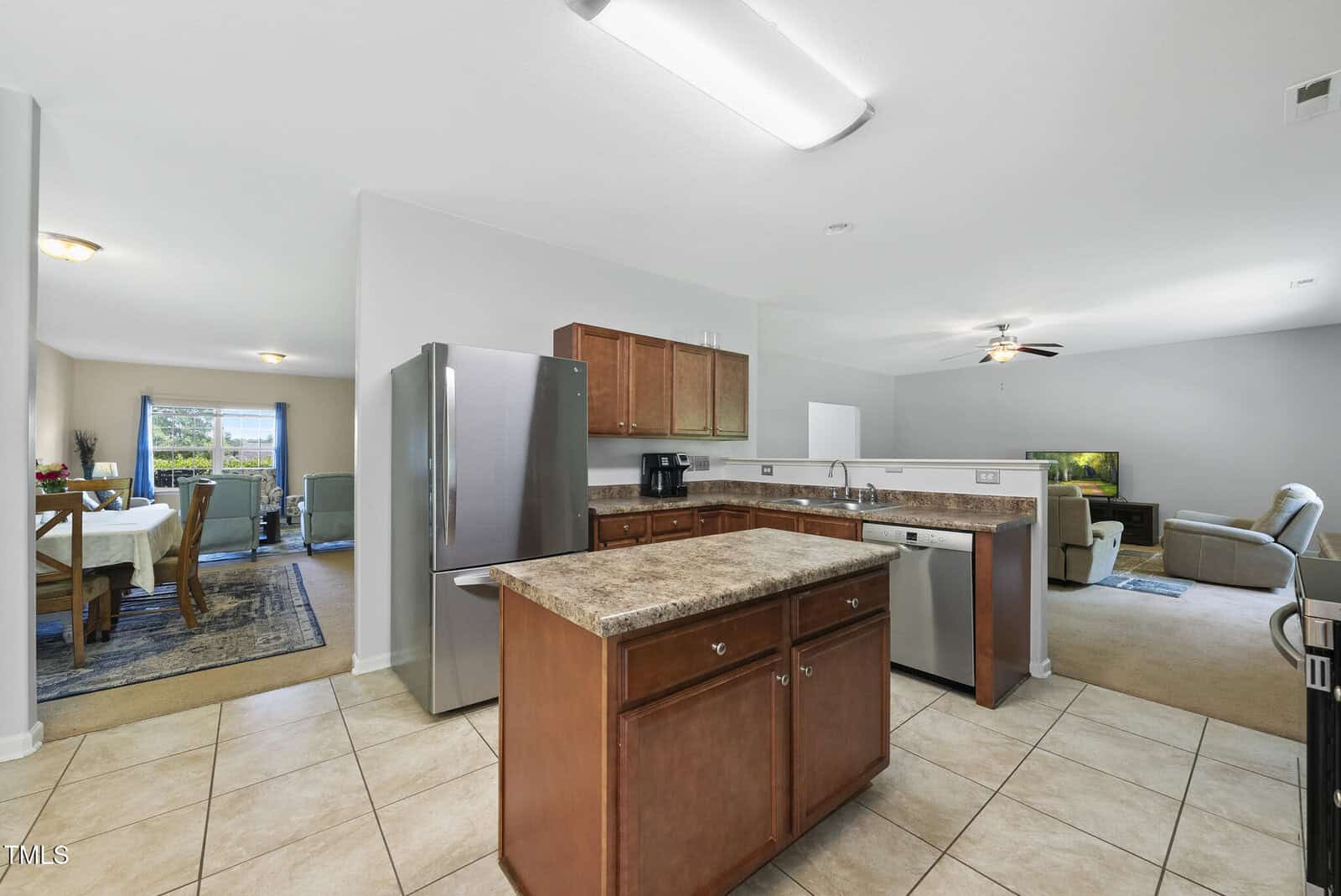A spacious open-concept kitchen with brown cabinets, an island, stainless steel appliances, and tile flooring, leading to a carpeted living area and a dining room with a table and chairs in the background.