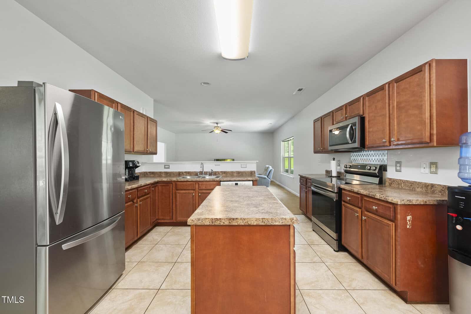 Spacious kitchen with brown cabinets, stainless steel appliances, a center island, tile flooring, and a water cooler. The room is well-lit with natural light and a ceiling light fixture.