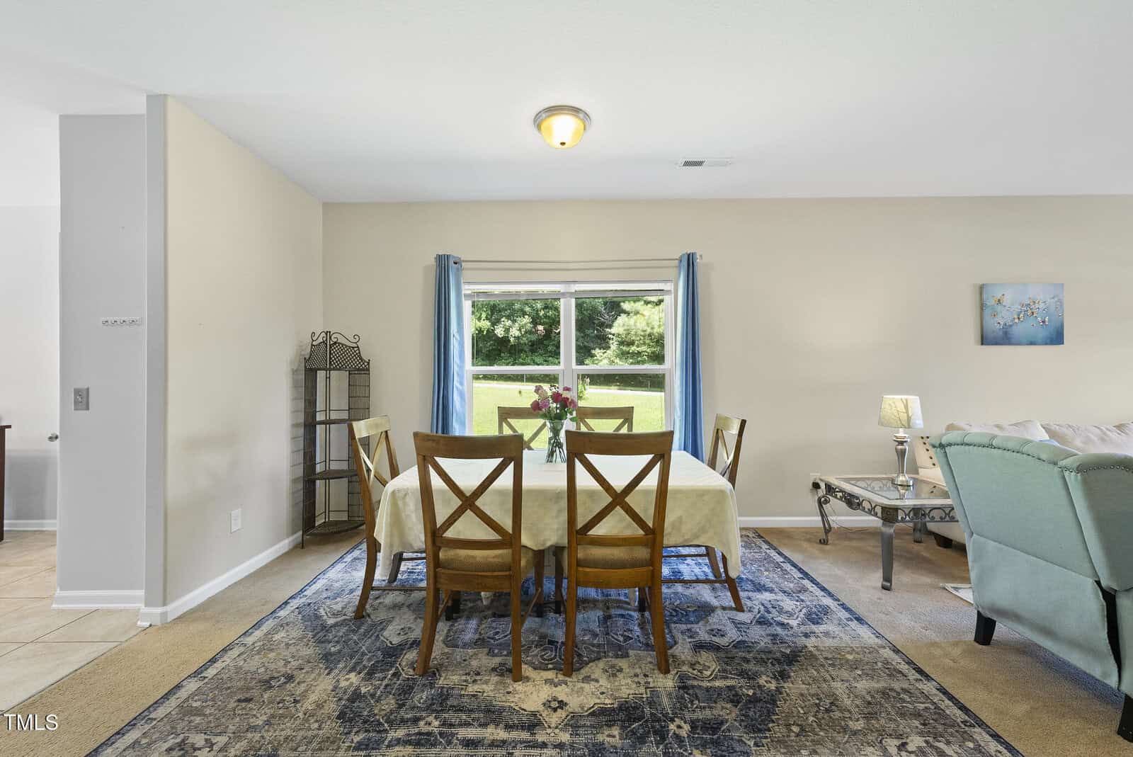 A dining area with a rectangular table covered by a cream tablecloth, four wooden chairs, a blue patterned rug, a vase with flowers, and a window with blue curtains overlooking greenery. Part of a living room is visible.