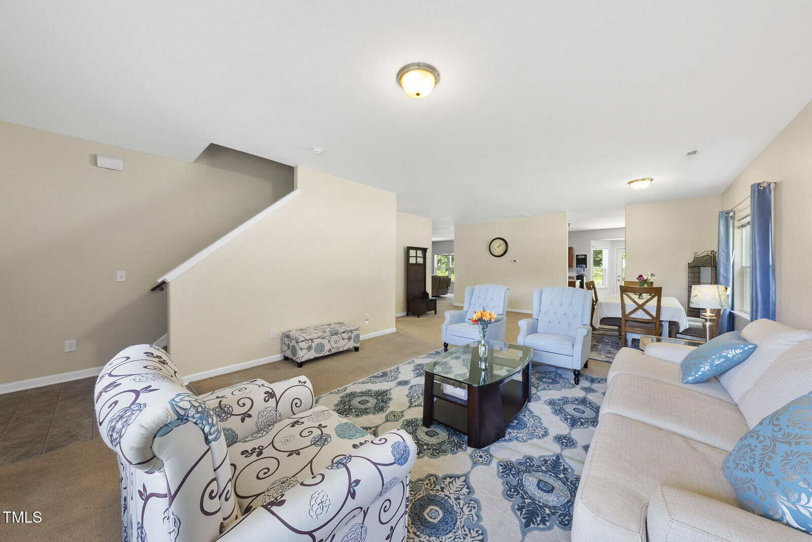 A spacious living room with beige walls, a light tan sofa, blue and floral-patterned armchairs, a glass coffee table, a rug with a blue pattern, and a staircase leading upstairs. The room is well-lit with ceiling lights.