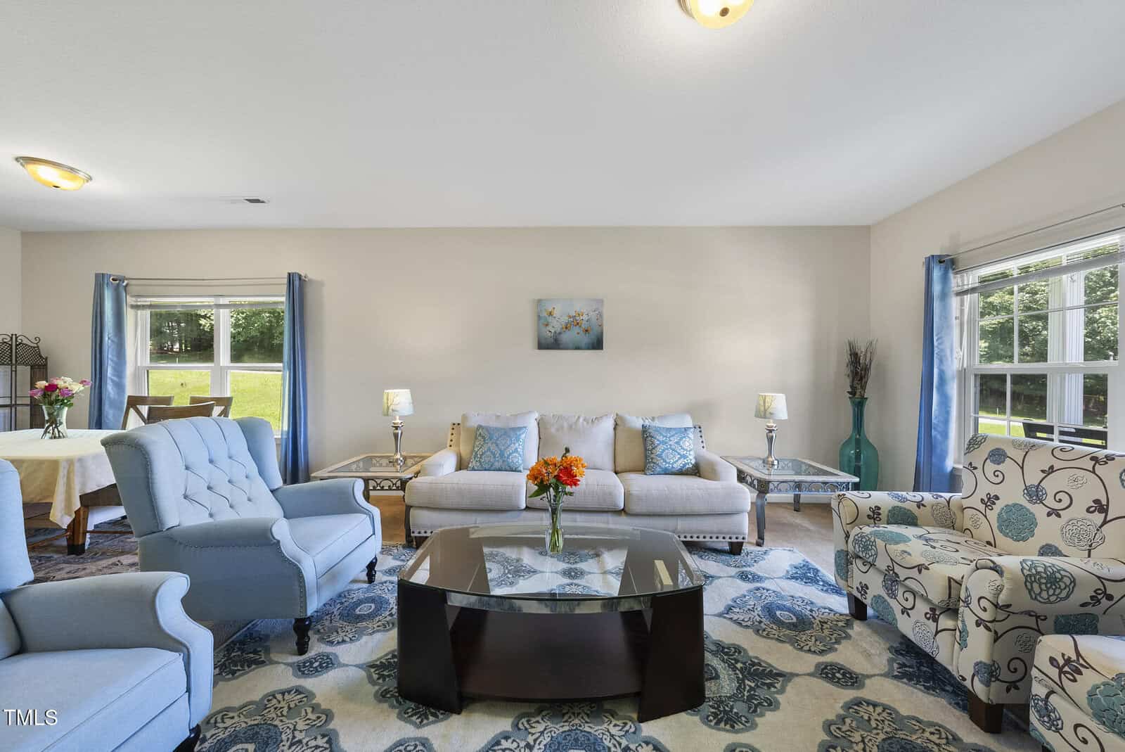 A living room with a beige sofa, two blue armchairs, a floral armchair, a glass coffee table with a vase of flowers, blue curtains, and large windows letting in natural light. A dining area is visible in the background.