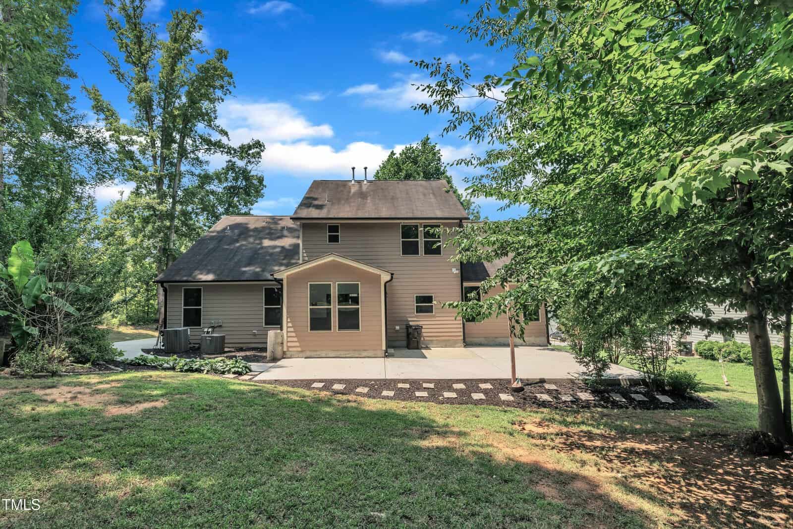 Two-story beige house with a dark roof, large windows, and a wide patio in the backyard, surrounded by green grass, trees, and landscaping under a bright blue sky with scattered clouds.