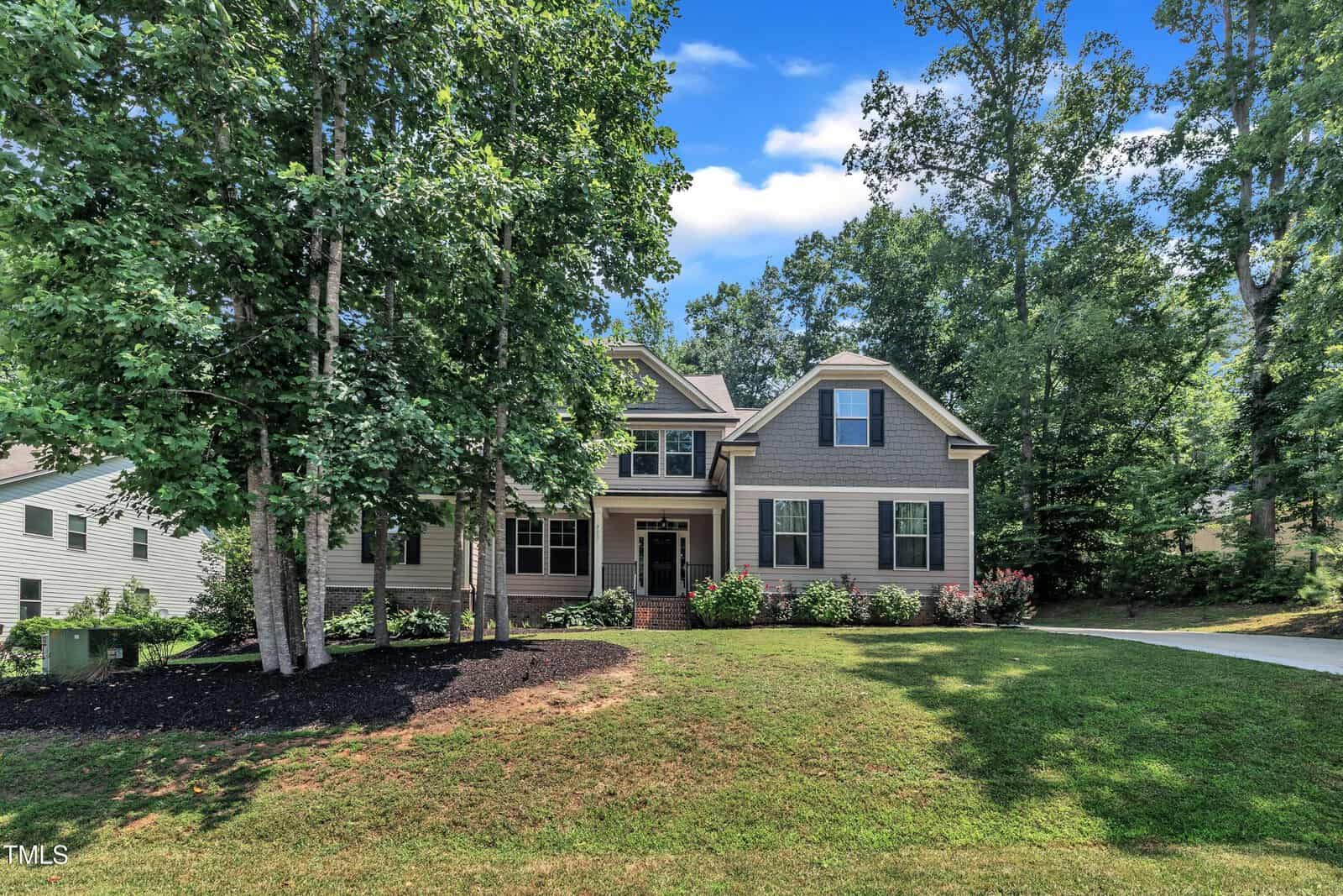 Two-story suburban house with gray siding and dark shutters, surrounded by trees and greenery. A neatly landscaped front yard with shrubs and a driveway is visible on a sunny day with a blue sky.