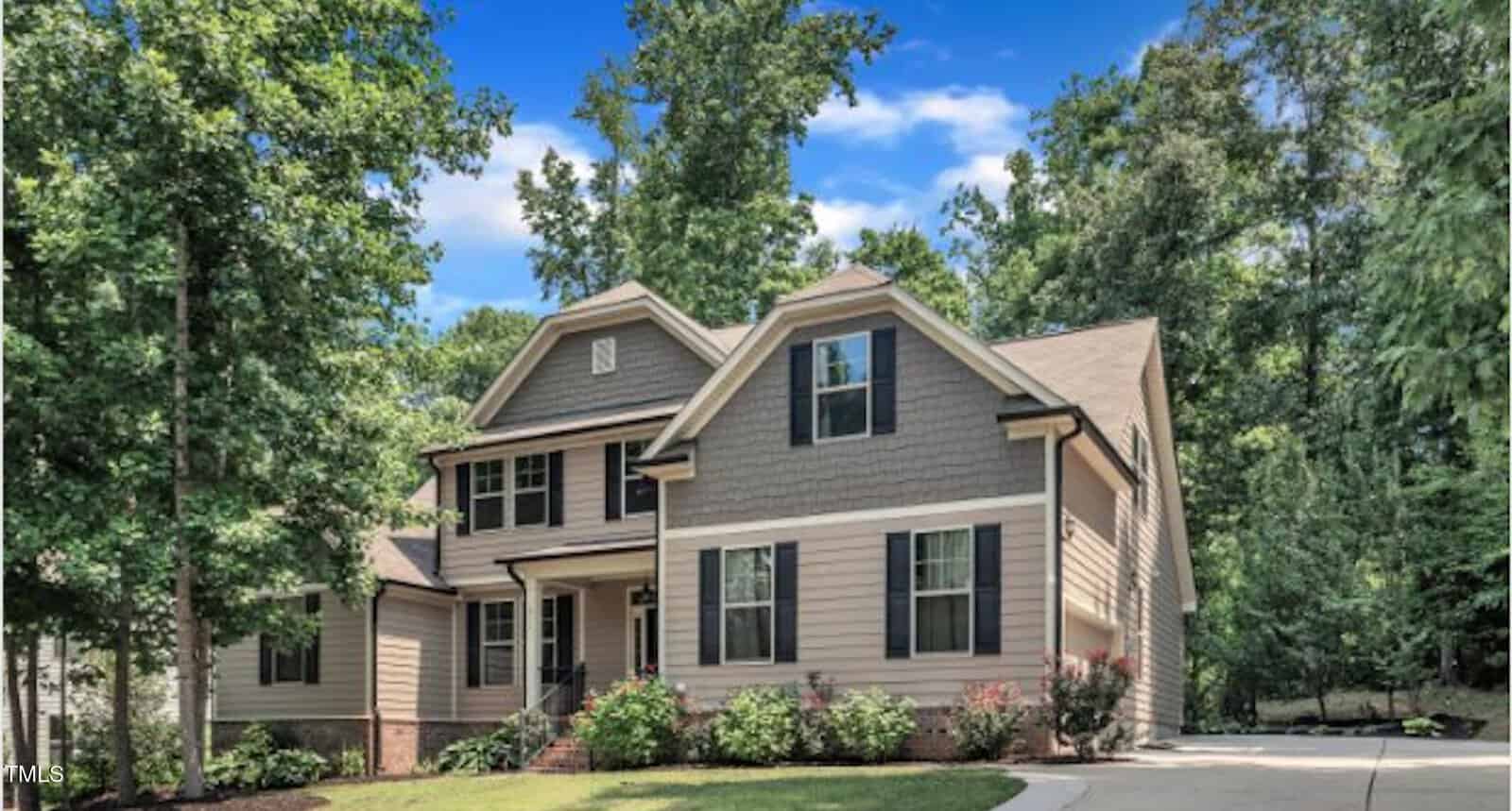 A beige two-story house with dark shutters and a gabled roof sits among tall trees, with a landscaped front yard and a curved driveway under a blue sky with scattered clouds.