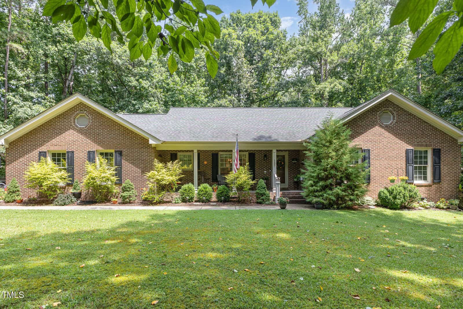 Single-story brick house with a dark roof, front porch, and columns, surrounded by neatly trimmed bushes, trees, and a green lawn, set against a wooded background under a clear sky.