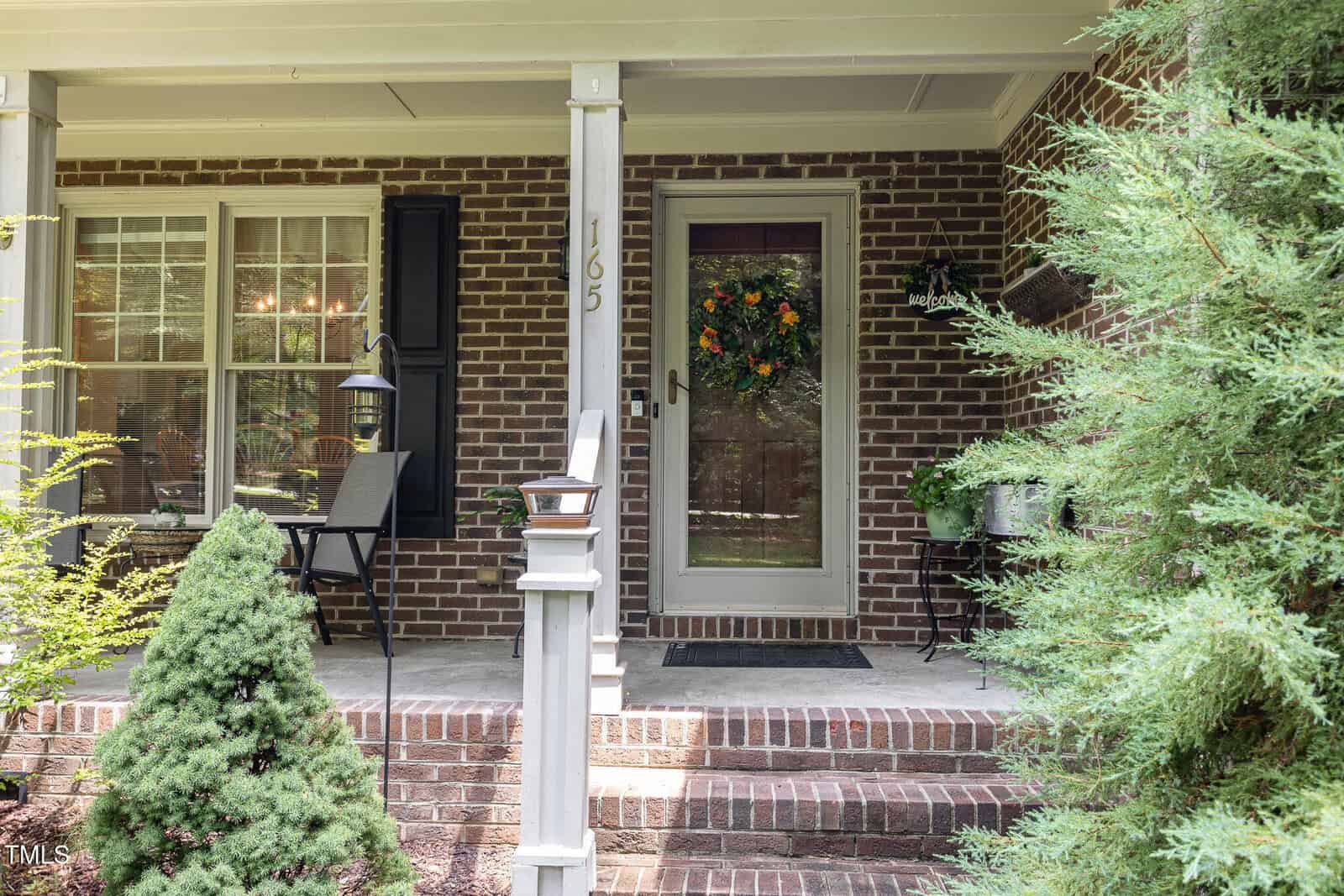 A brick house front porch with steps, a glass door decorated with a floral wreath, two chairs, a small table, potted plants, and greenery around the entrance.