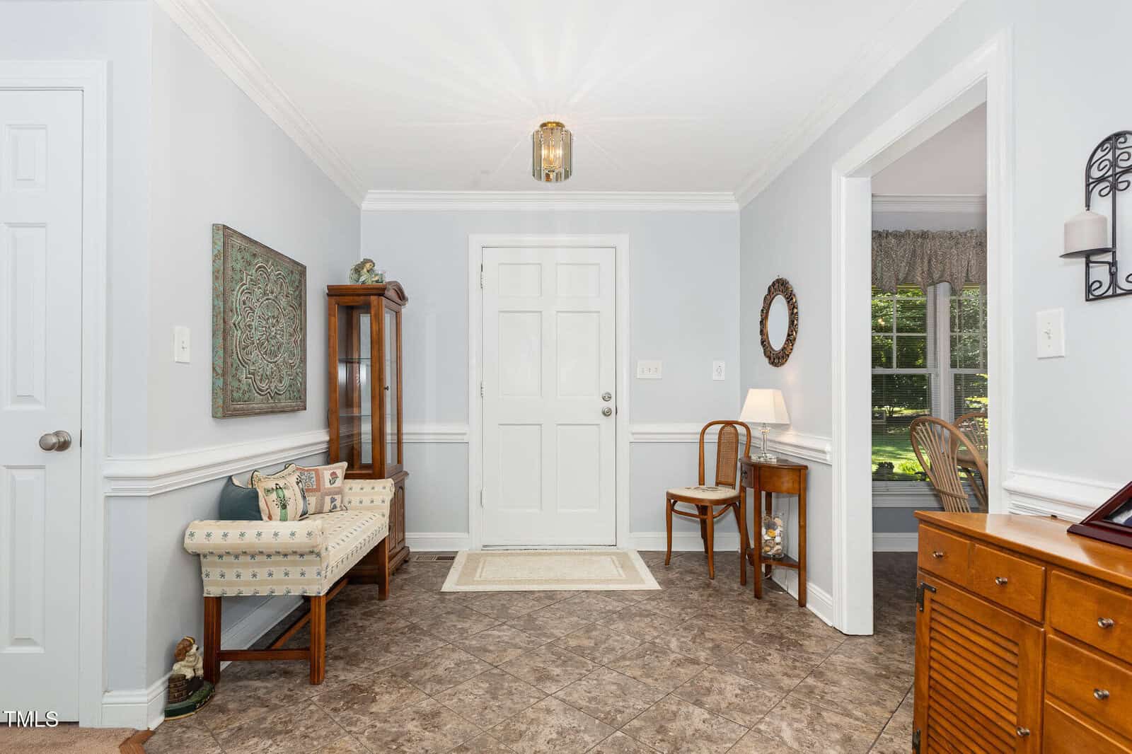 A bright, tidy entryway with white walls, tile floor, a decorative bench, display cabinet, small wooden tables, chairs, wall art, and a window overlooking a green yard.