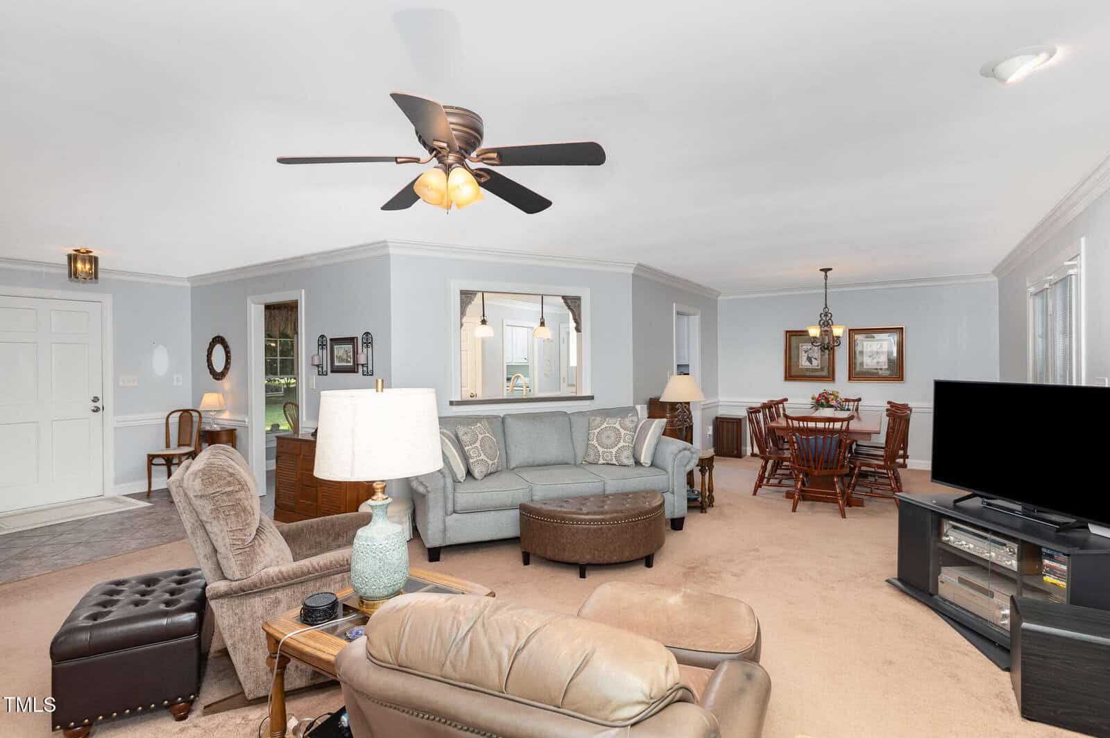 A spacious living and dining area with neutral walls, beige carpet, two armchairs, a sectional sofa, a ceiling fan, a round ottoman, and a flat-screen TV. The dining table seats six under a chandelier.
