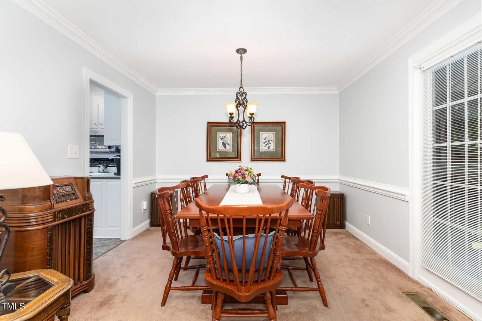 A dining room with a wooden table and eight chairs, a chandelier overhead, two framed pictures on the back wall, beige carpet, and a window with blinds to the right. A vintage radio stands in the left foreground.