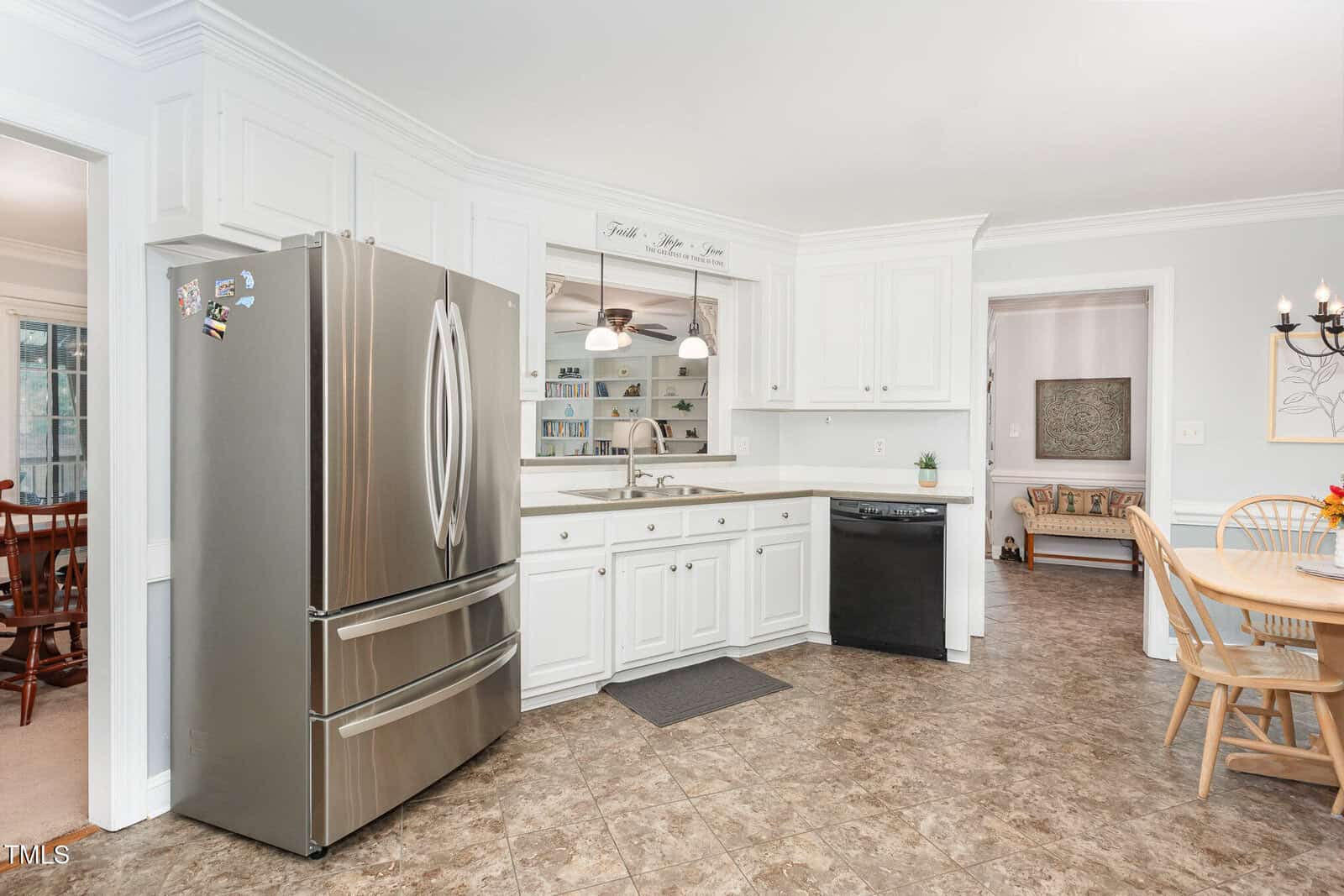 A kitchen with white cabinets, stainless steel refrigerator, black dishwasher, double sink, beige tiled floor, and adjoining dining and living areas visible through open doorways. Ceiling-mounted lights above the sink.