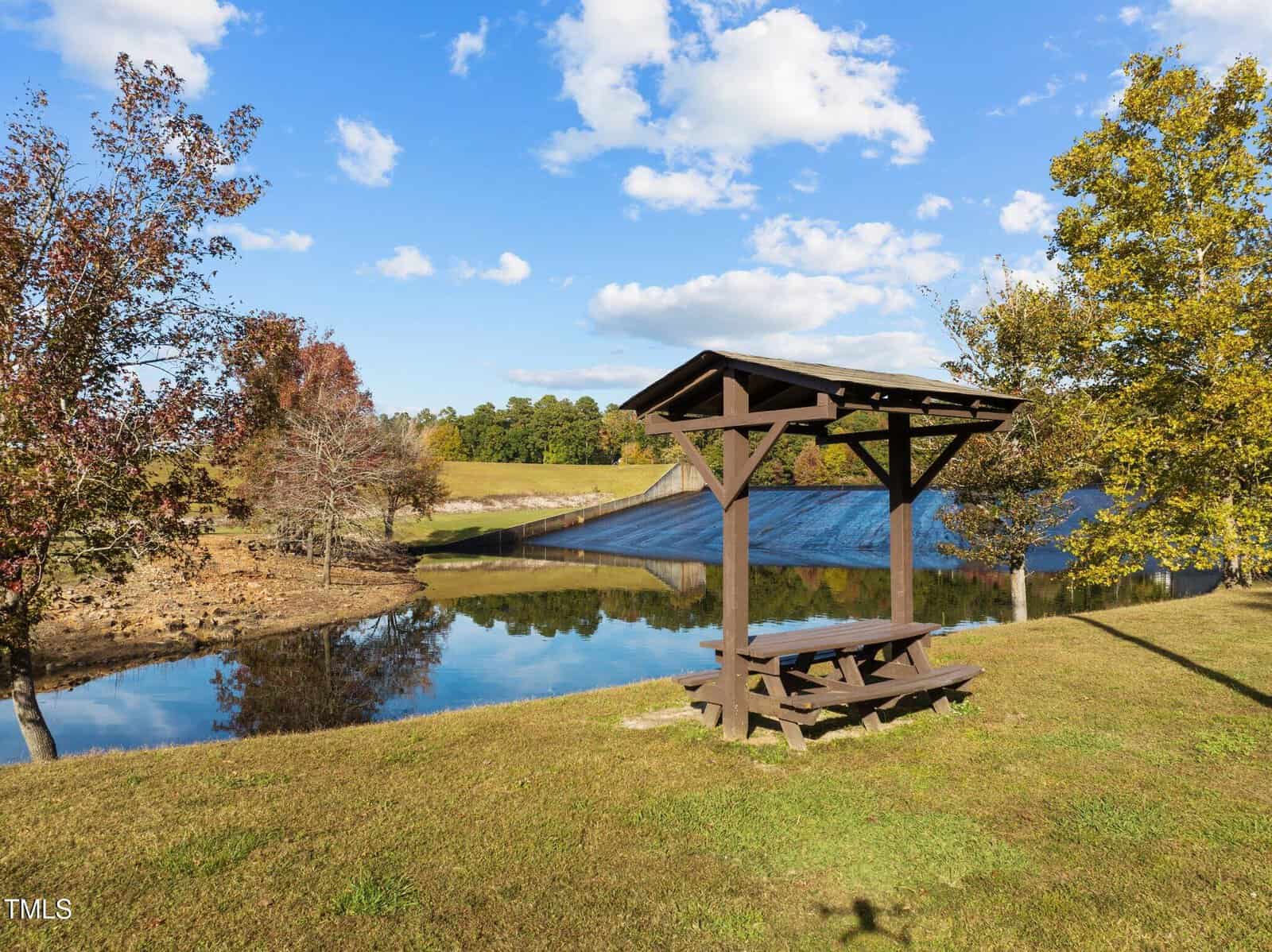 A wooden picnic table with a shelter sits on grass by a calm pond, reflecting trees and a clear blue sky with scattered clouds. Trees, some with autumn foliage, surround the area.