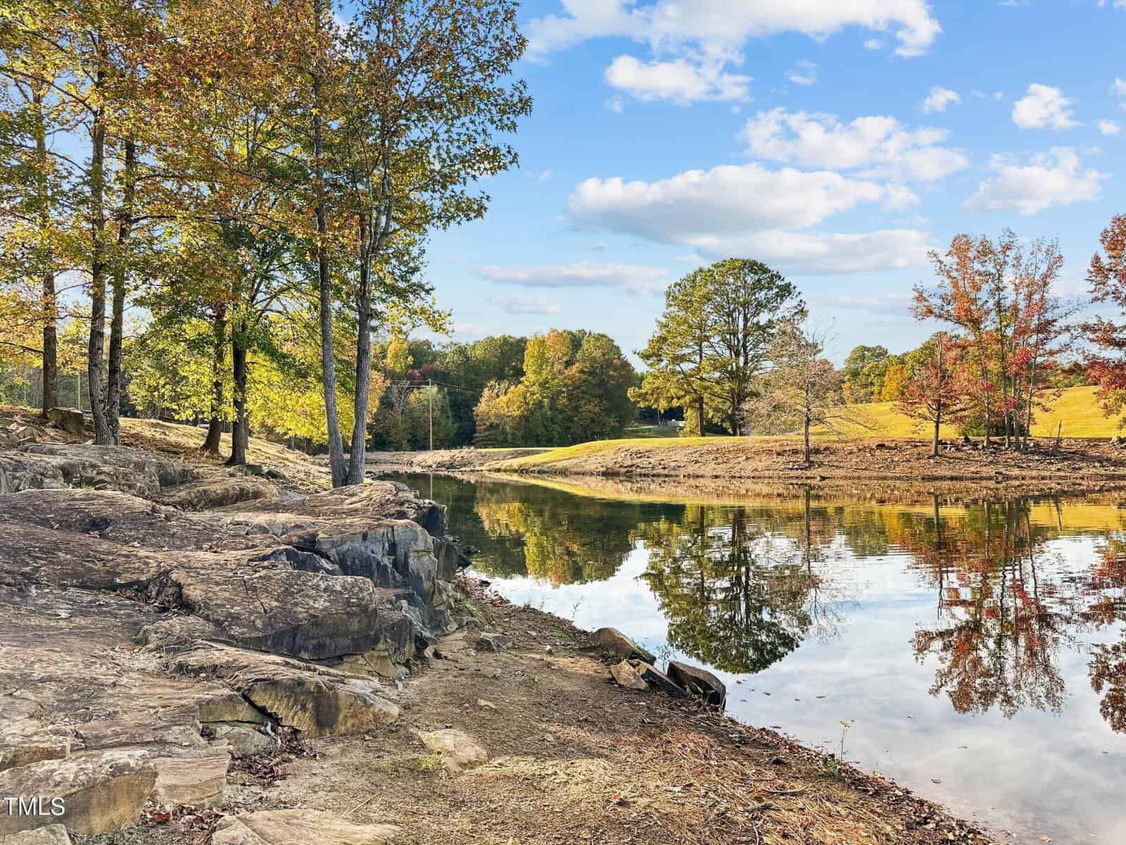 A calm pond reflects trees with autumn foliage and a blue sky with scattered clouds. Rocky ground and fallen leaves cover the shoreline, and the landscape extends into grassy, tree-dotted slopes in the background.