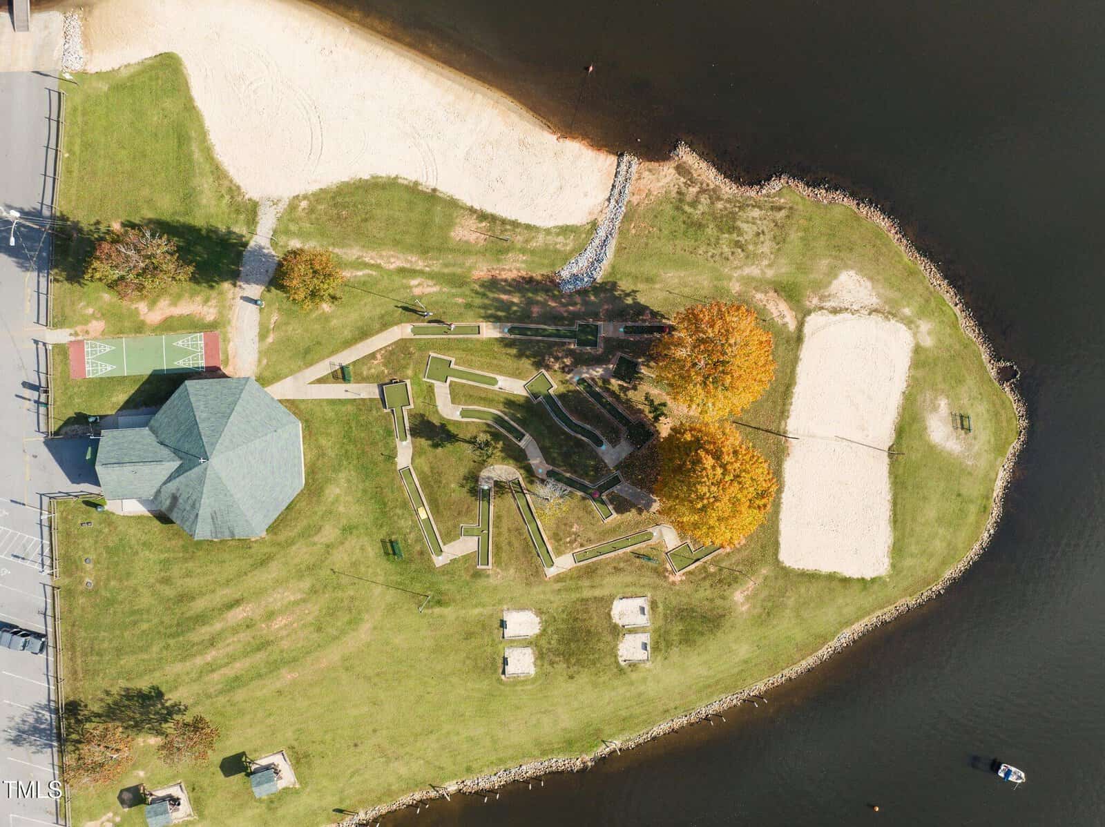 Aerial view of a small lakeside park with a pavilion, basketball court, walking paths, picnic tables, two sand areas, and trees with autumn foliage, bordered by grass and water. A small boat is visible on the lake.