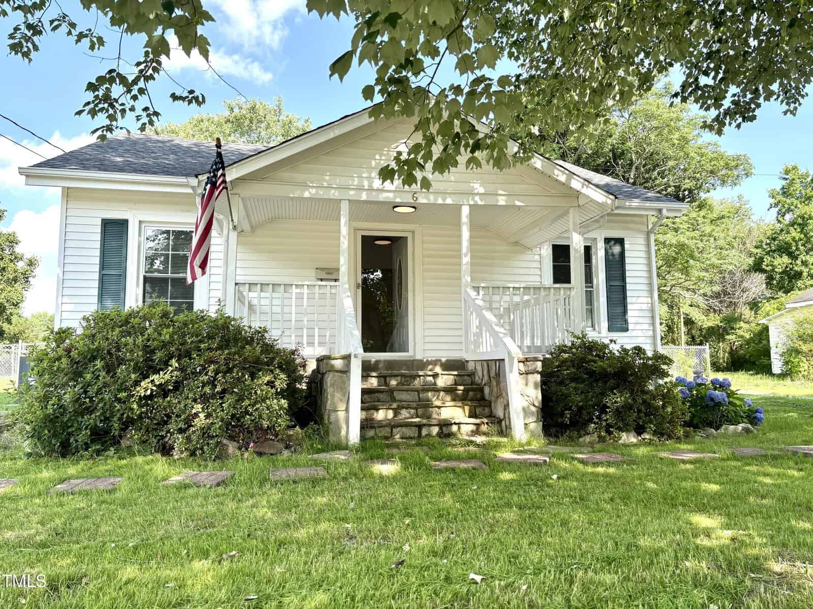 Charming single-story white house with a covered front porch, steps, and railing located at 6 First Street, Franklinton. An American flag hangs on the left, welcoming you home. Lush green lawn, shrubs, and trees surround the house on this bright, sunny day with a clear sky.