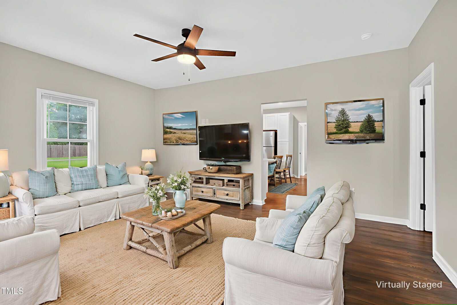 A bright living room with beige walls, two white sofas, a wooden coffee table, a TV on a stand, wall art, wooden floors, and a ceiling fan. A doorway leads to the kitchen. “Virtually Staged” text is in the corner.