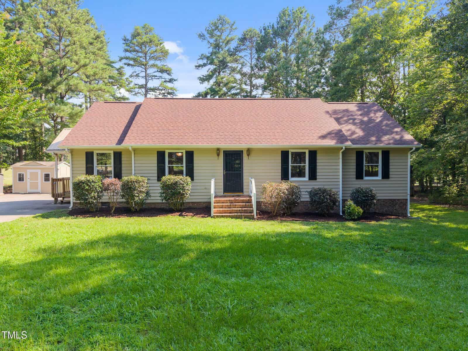 A single-story house with beige siding, dark shutters, and a red roof sits on a well-kept lawn at 2856 Nutbush Road, Henderson. Bushes line the front and trees fill the background, with a small outbuilding visible on the left.