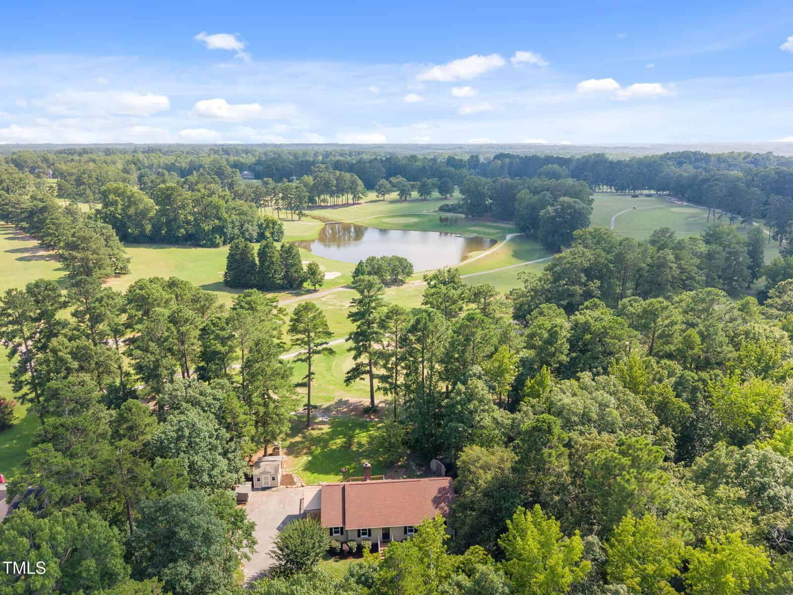 Aerial view of a house surrounded by trees with a golf course, pond, and open grassy areas in the background under a blue sky with scattered clouds.