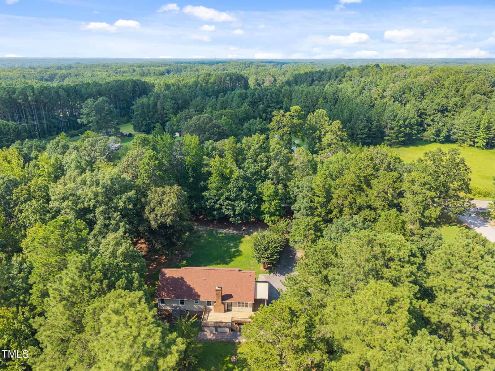 Aerial view of a two-story house with a brown roof surrounded by dense green trees, open grassy areas, and a forested landscape under a blue sky with scattered clouds.