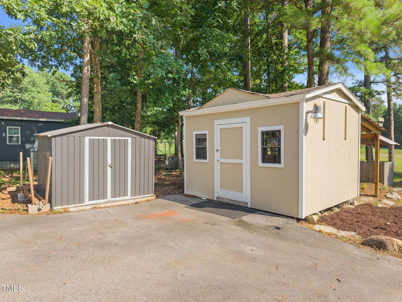 A small beige wooden shed with windows and a white door is next to a gray metal storage shed on a paved area, with trees and another dark-colored building in the background.