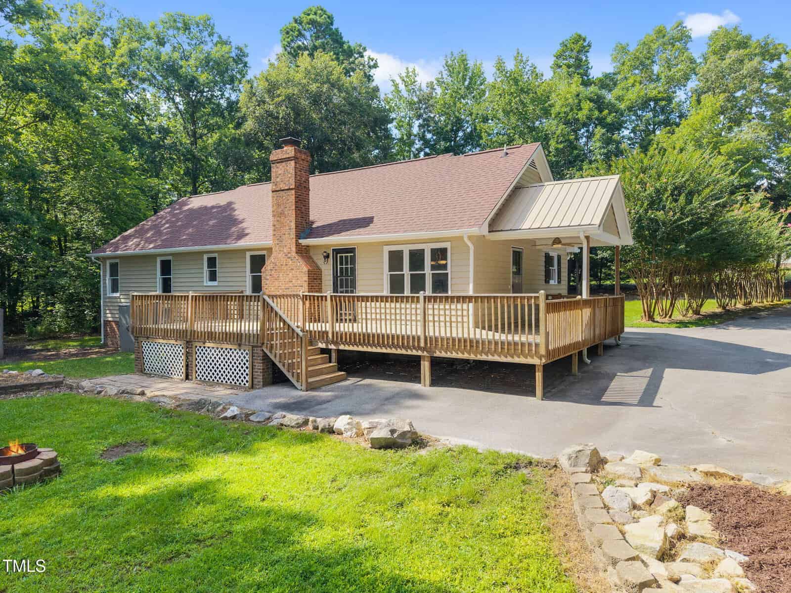 A single-story beige house with a brown roof, large front wooden deck, and brick chimney, surrounded by trees, with a paved driveway and grassy yard in the foreground.